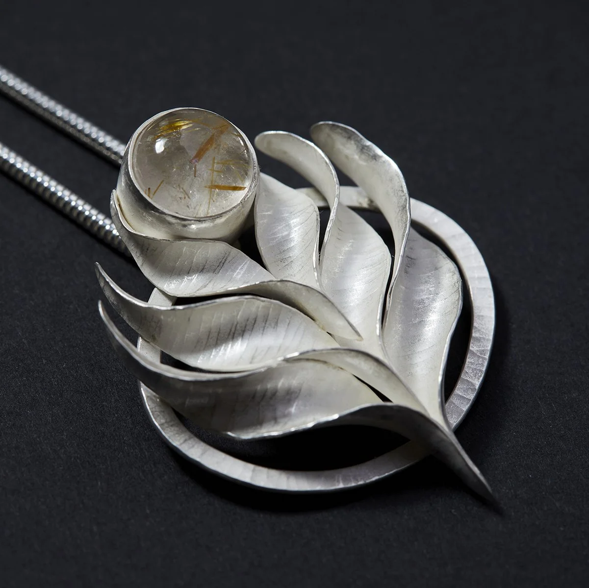A silver floral-shaped pendant with a round rutilated quartz gemstone (clear but shot through with gold coloured threads) at the top, on a 1.9mm silver snake chain, on a black background.