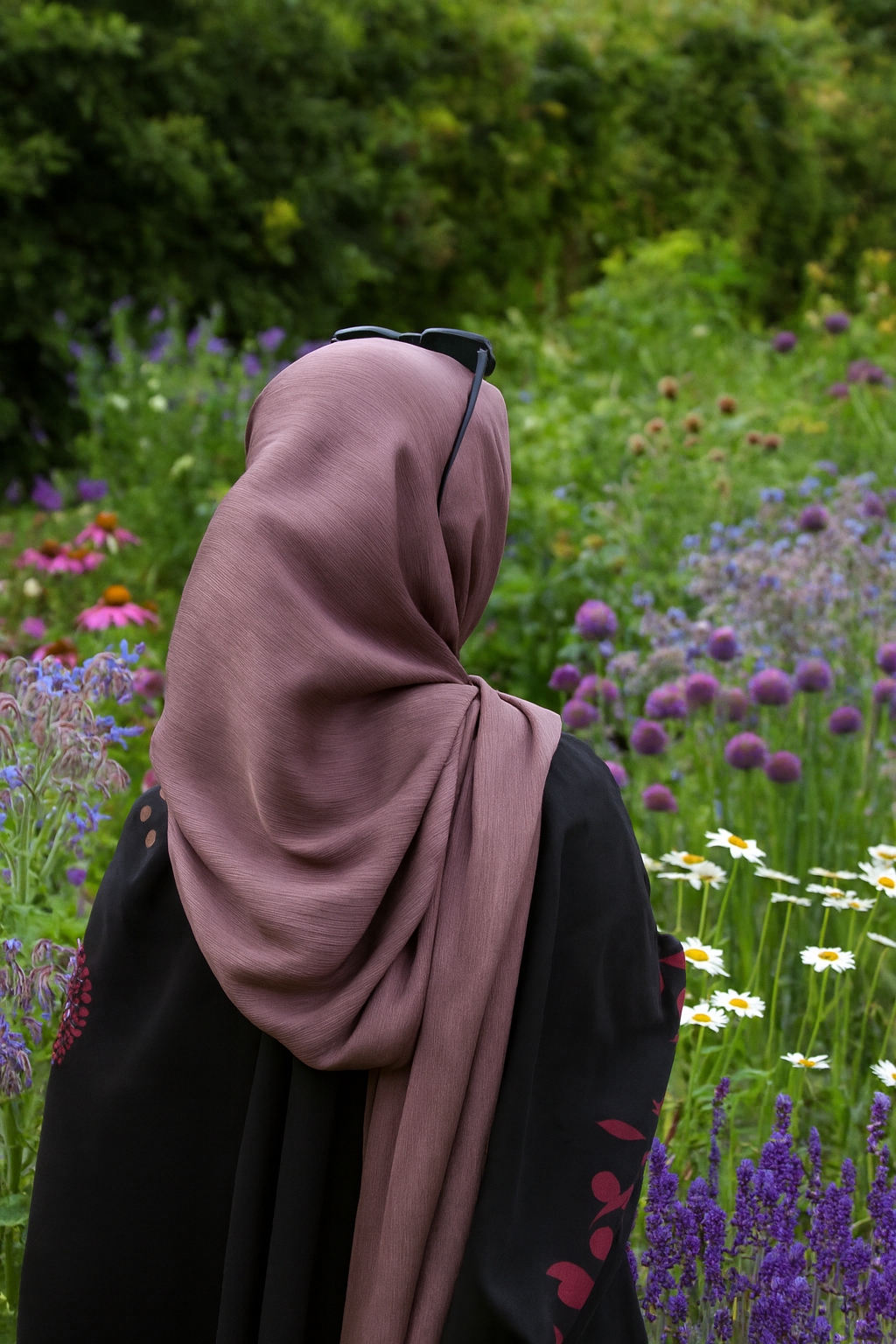 A woman wearing a brown headscarf and glasses on her head stands in a lush garden with colorful flowers.