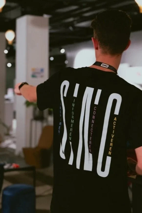 Een man met een zwart T-shirt dat de woorden 'SHIC' en enkele Nederlandse woorden erop heeft, wijzend met zijn arm in een binnenruimte met een donkere plafond en wat meubels op de achtergrond.