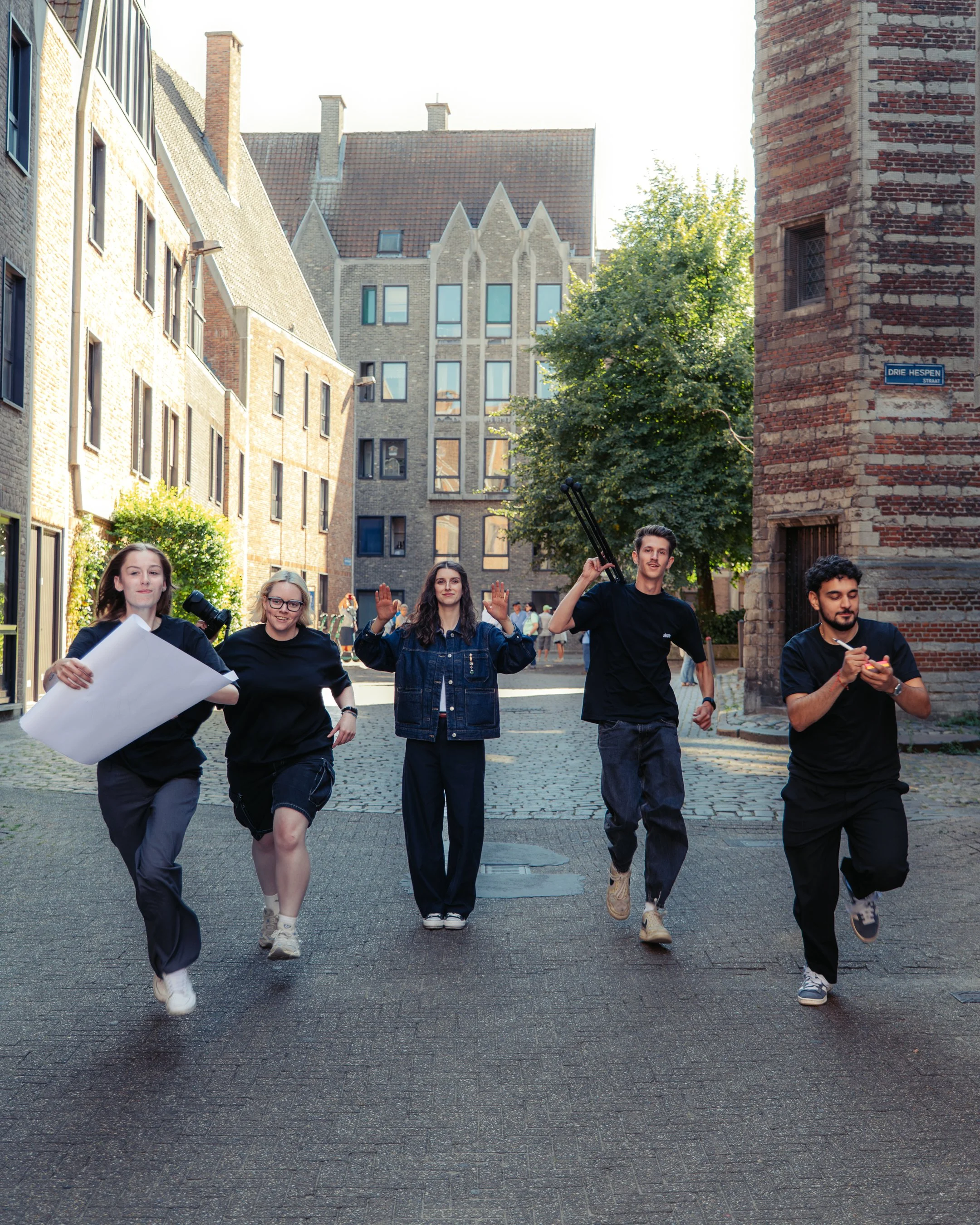 Een groep jonge mensen die wandelen op een straat in een oude wijk met klinkers, omringd door historische gebouwen en een boom, onder een heldere hemel.