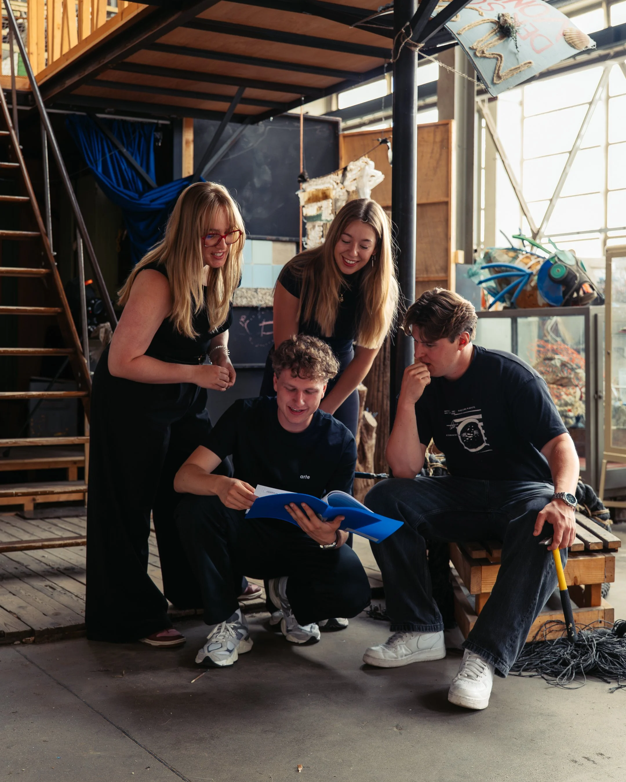 Vijf jonge mensen zitten en staan samen op een houten platform onder een industriële structuur, ze kijken naar een boek of document dat één persoon vasthoudt. Ze lijken betrokken en lachen.