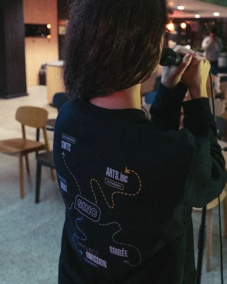 Persoon met kort bruin haar die met een microfoon spreekt in een indoor ruimte, zittend op een stoel, met lege stoelen op de achtergrond.