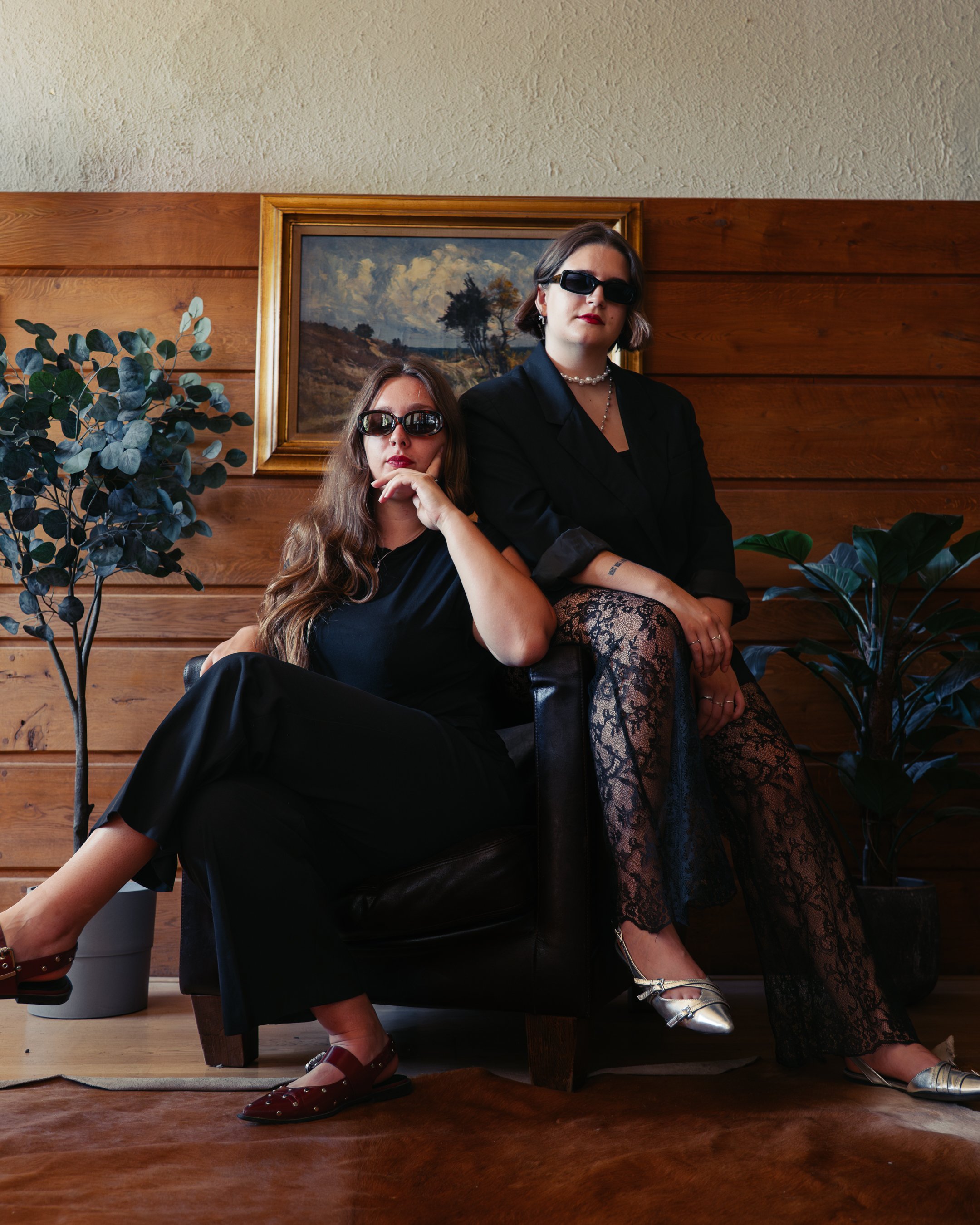 Twee vrouwen met donkere zonnebrillen en strakke kleren zitten samen in een houten interieur met een landschapsschilderij en groene planten op de achtergrond.
