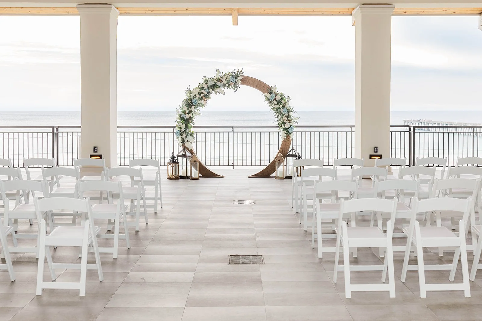 Setup for a wedding ceremony on a covered outdoor patio overlooking the ocean, featuring white folding chairs arranged in rows, a circular floral arch, lanterns, and a railing with a pier in the distance.
