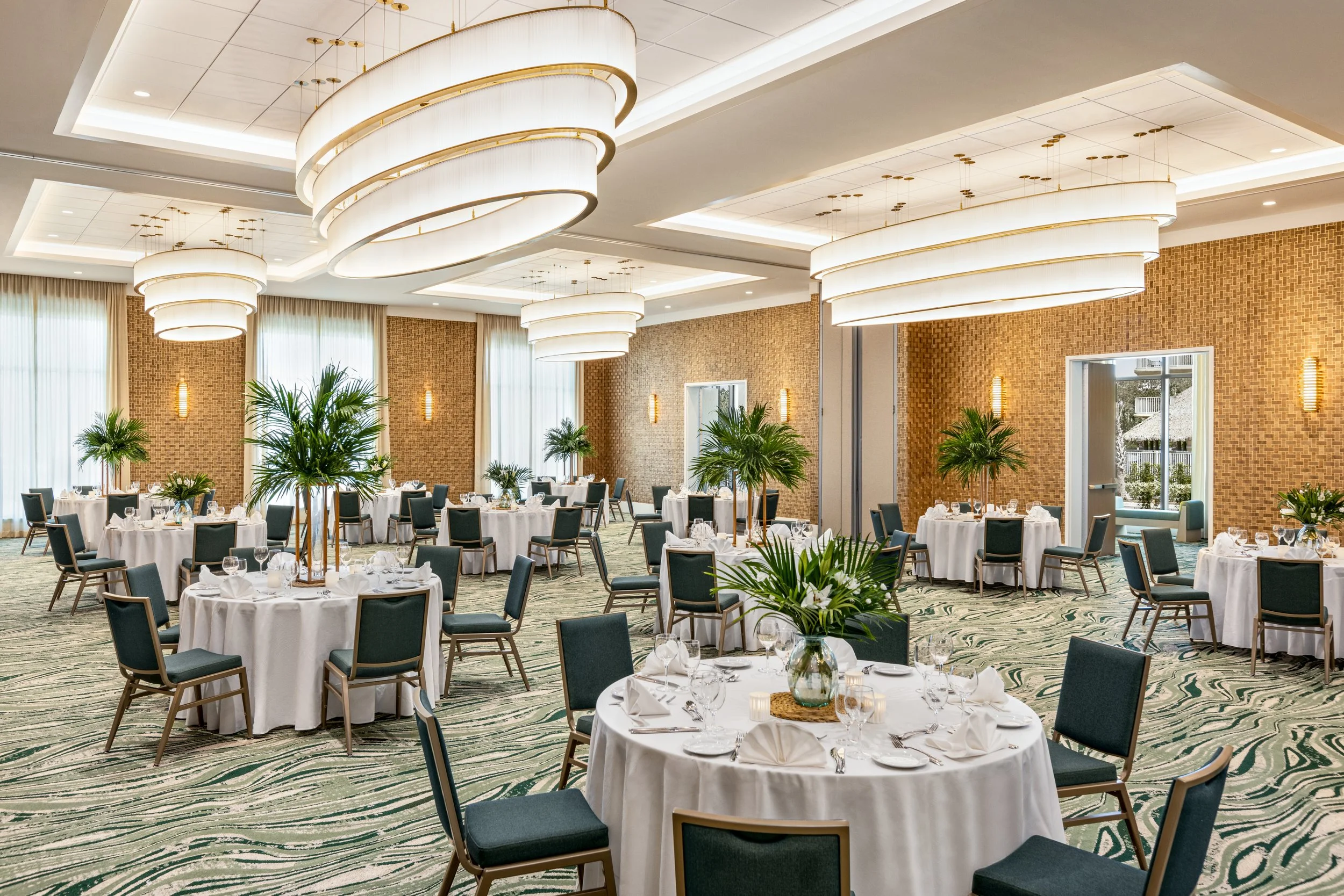 Elegant banquet hall with round tables covered in white tablecloths and surrounded by dark green chairs, decorated with glassware and folded napkins. Large modern circular chandeliers hang from the ceiling, with sunlight coming through tall windows draped with beige curtains. The walls are decorated with textured orange panels and wall-mounted lights, and there are green potted plants adding a natural touch to the space.