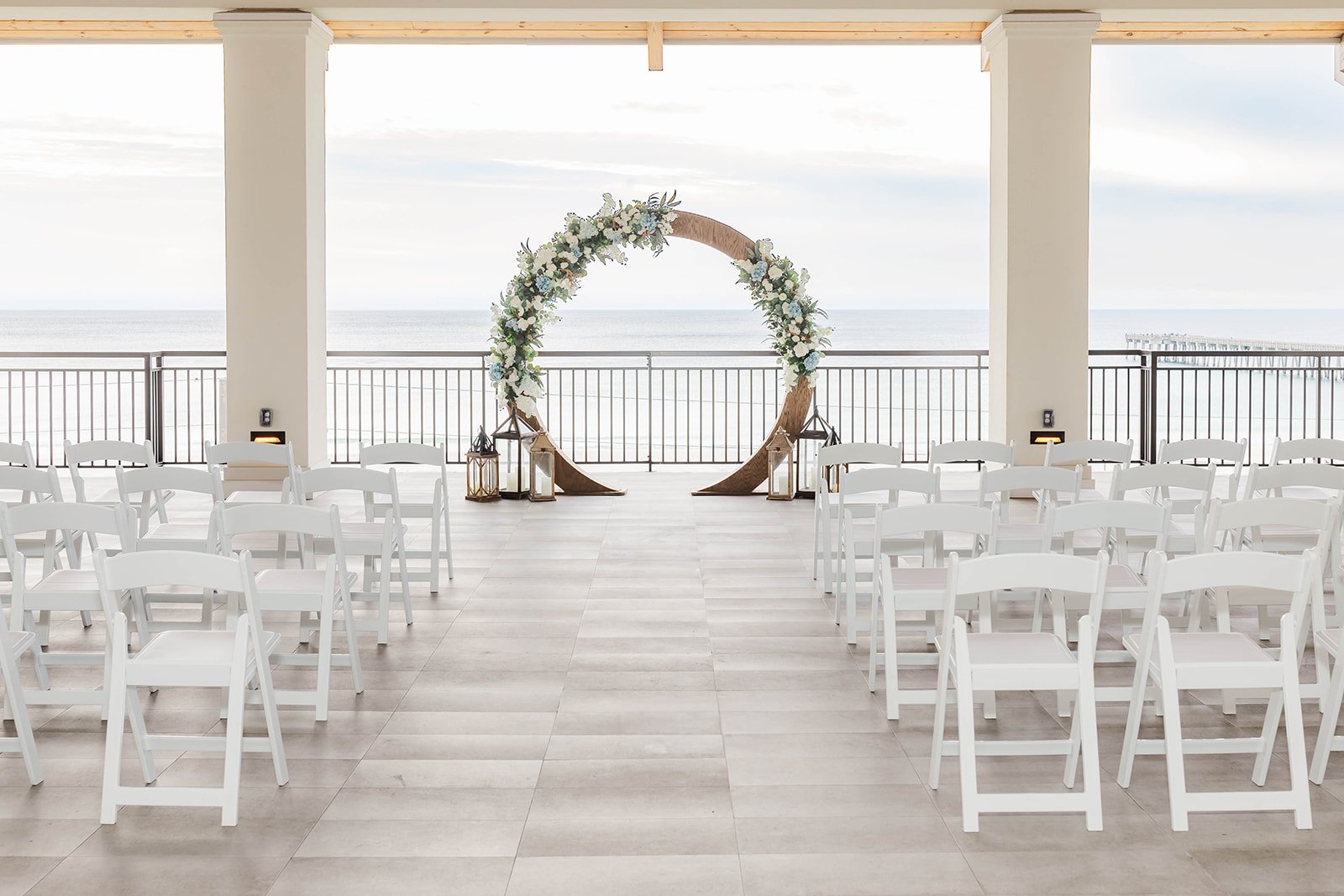 Setup for a wedding ceremony on a covered outdoor patio overlooking the ocean, featuring white folding chairs arranged in rows, a circular floral arch, lanterns, and a railing with a pier in the distance.