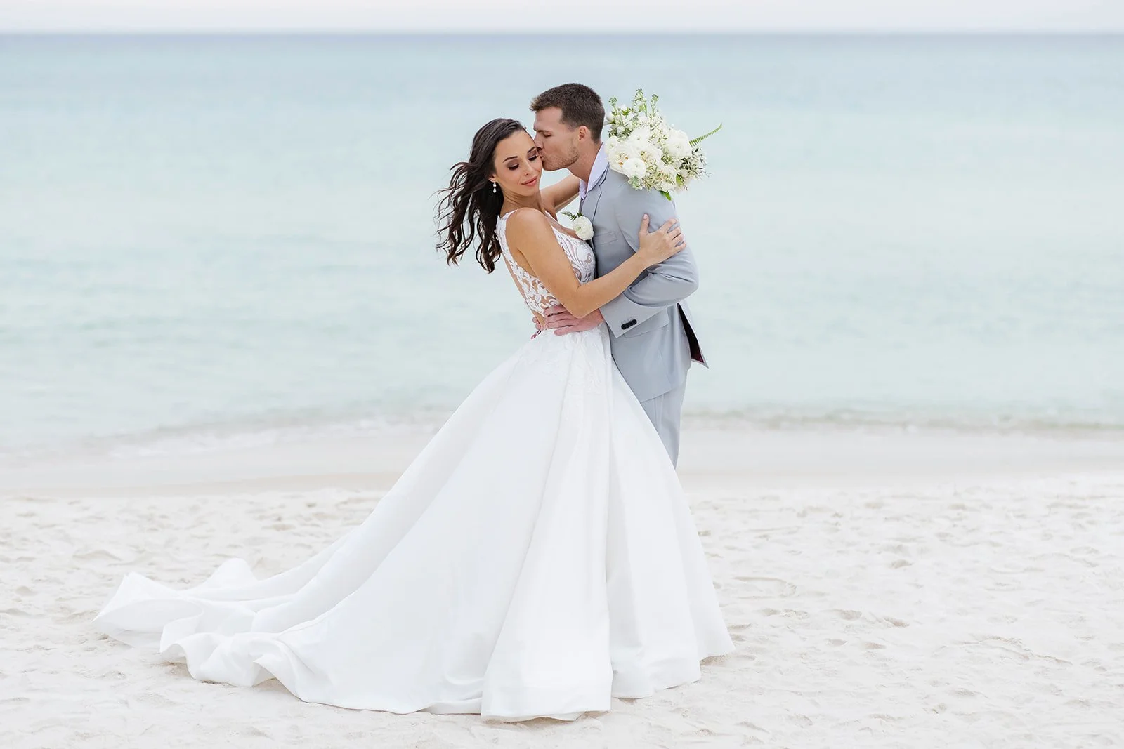 A groom kisses his bride on the cheek in front of clear blue water and white sand on the beach.