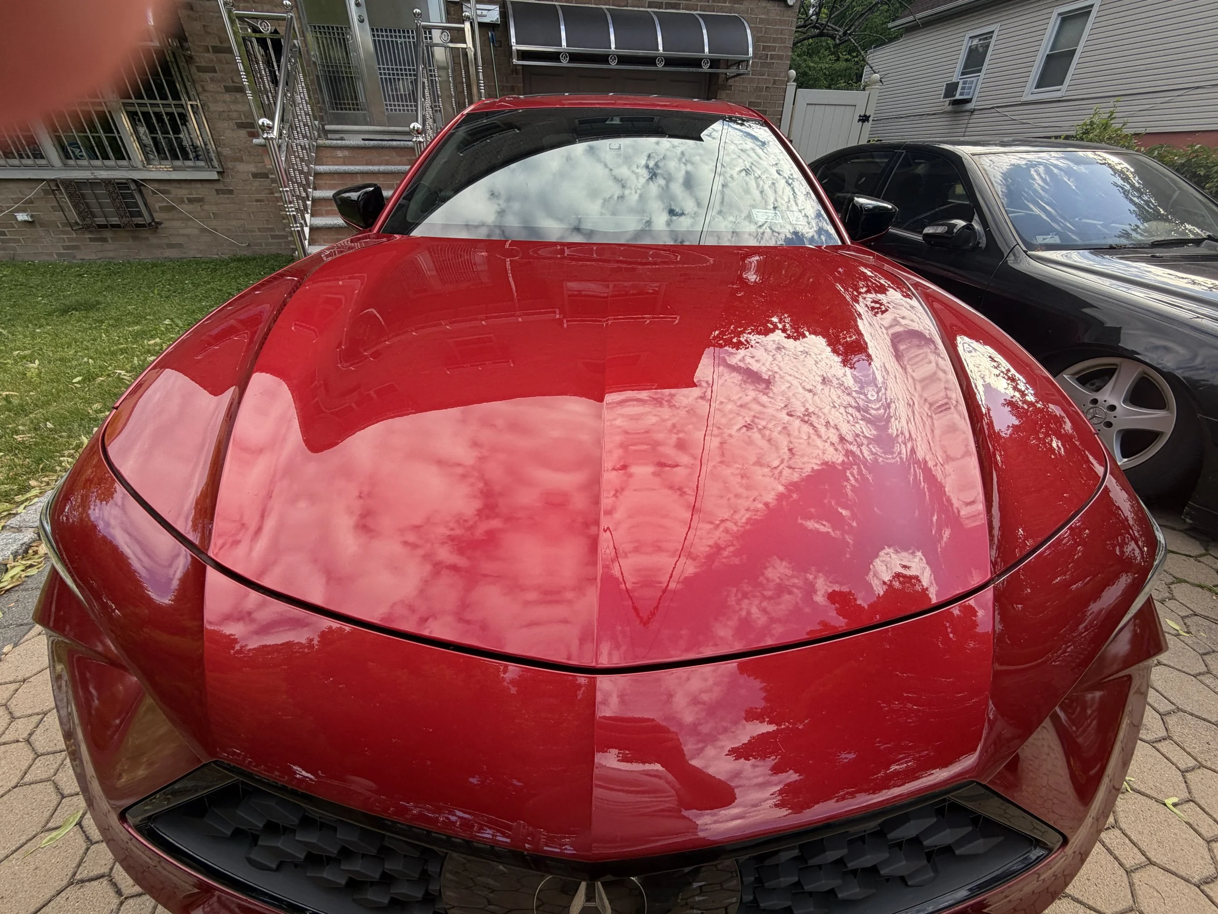 A red sports car is parked next to a black car on a driveway, reflecting clouds and sky on its shiny hood and windshield.