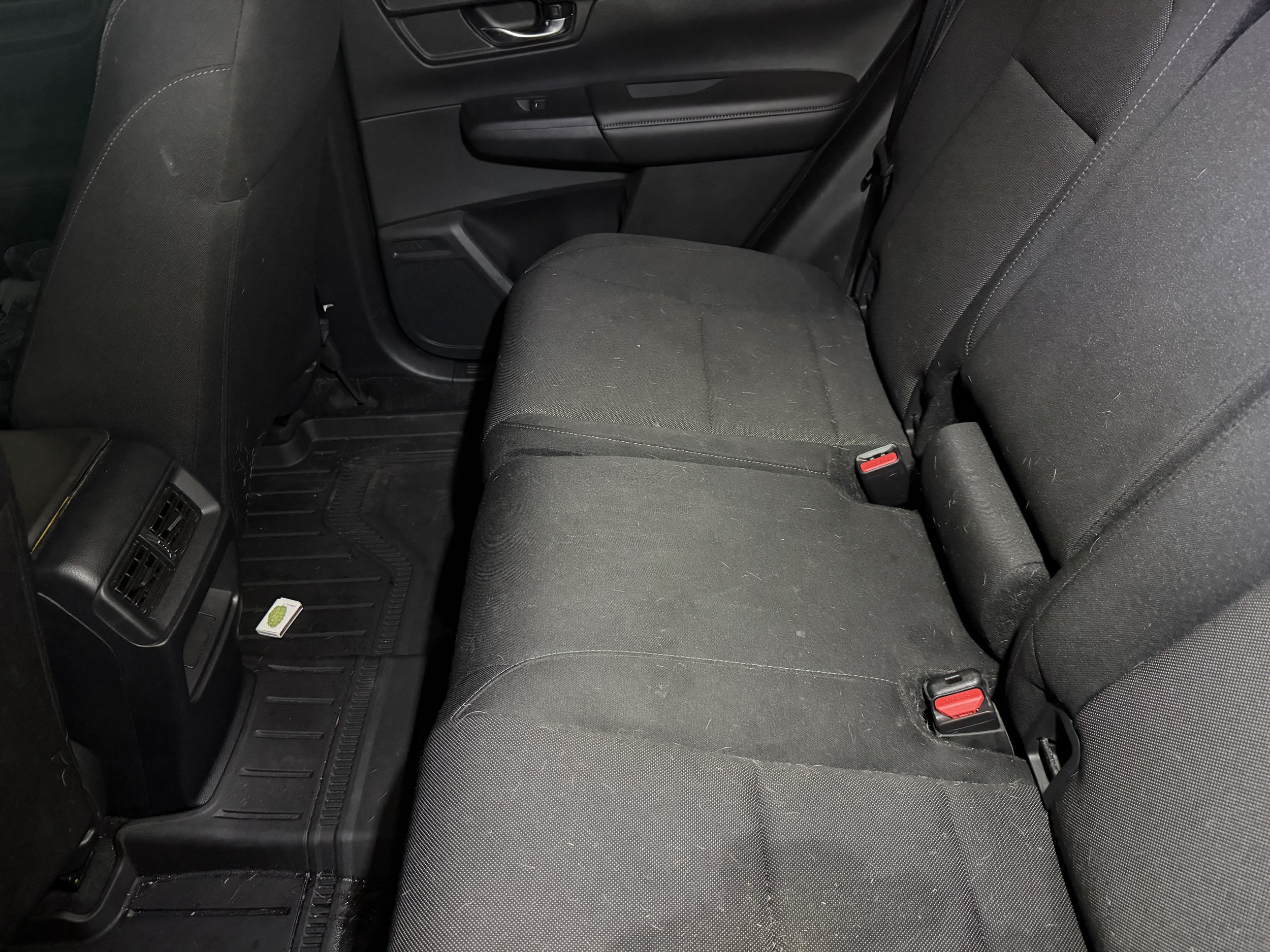 Backseat of a car with black fabric seats, plastic door panels, a floor mat, and a small green and white pack of gum on the floor.