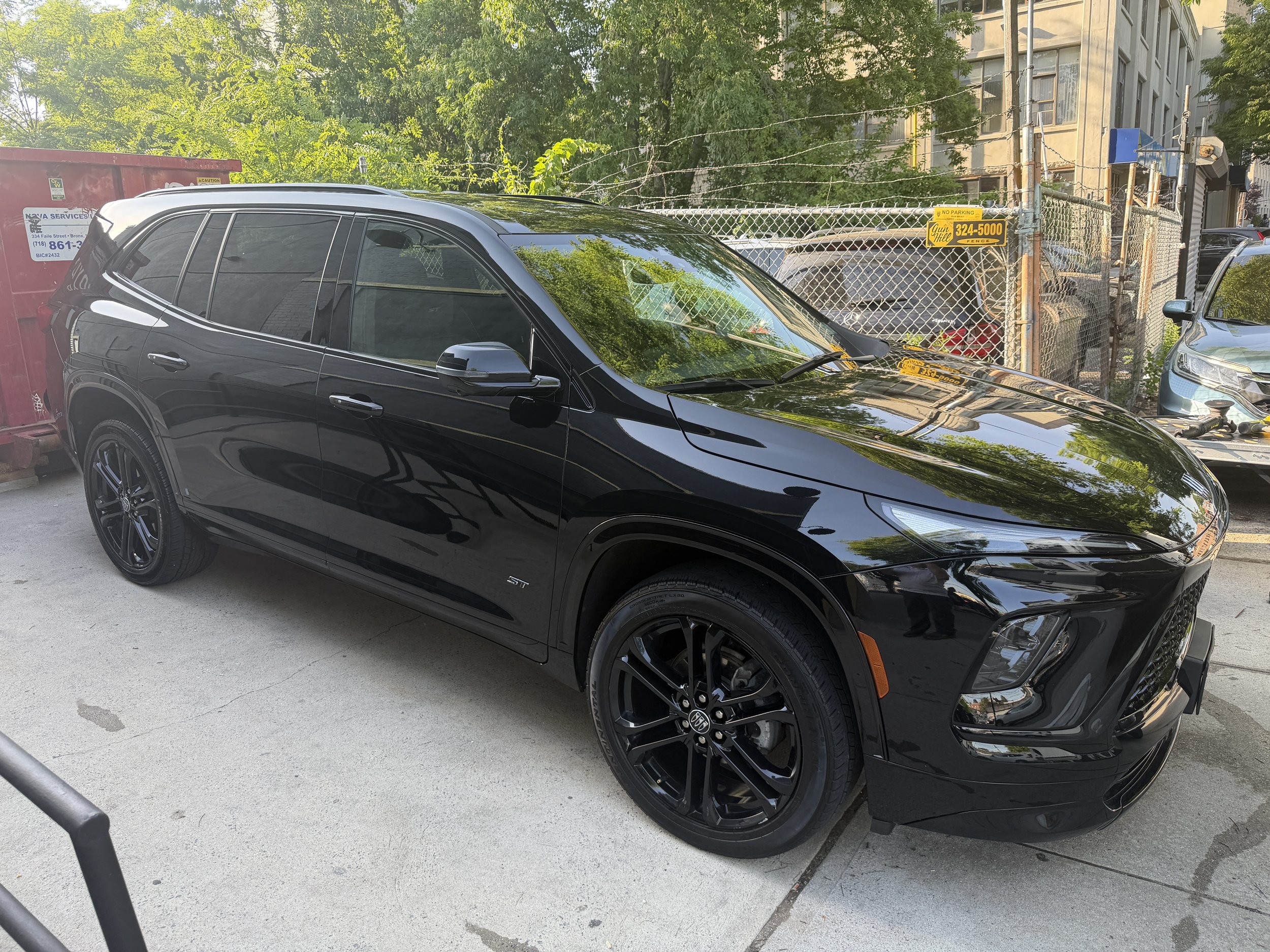 A row of cars parked along a street, featuring a black SUV in the foreground and a part of another vehicle to the right. There are trees and buildings in the background.
