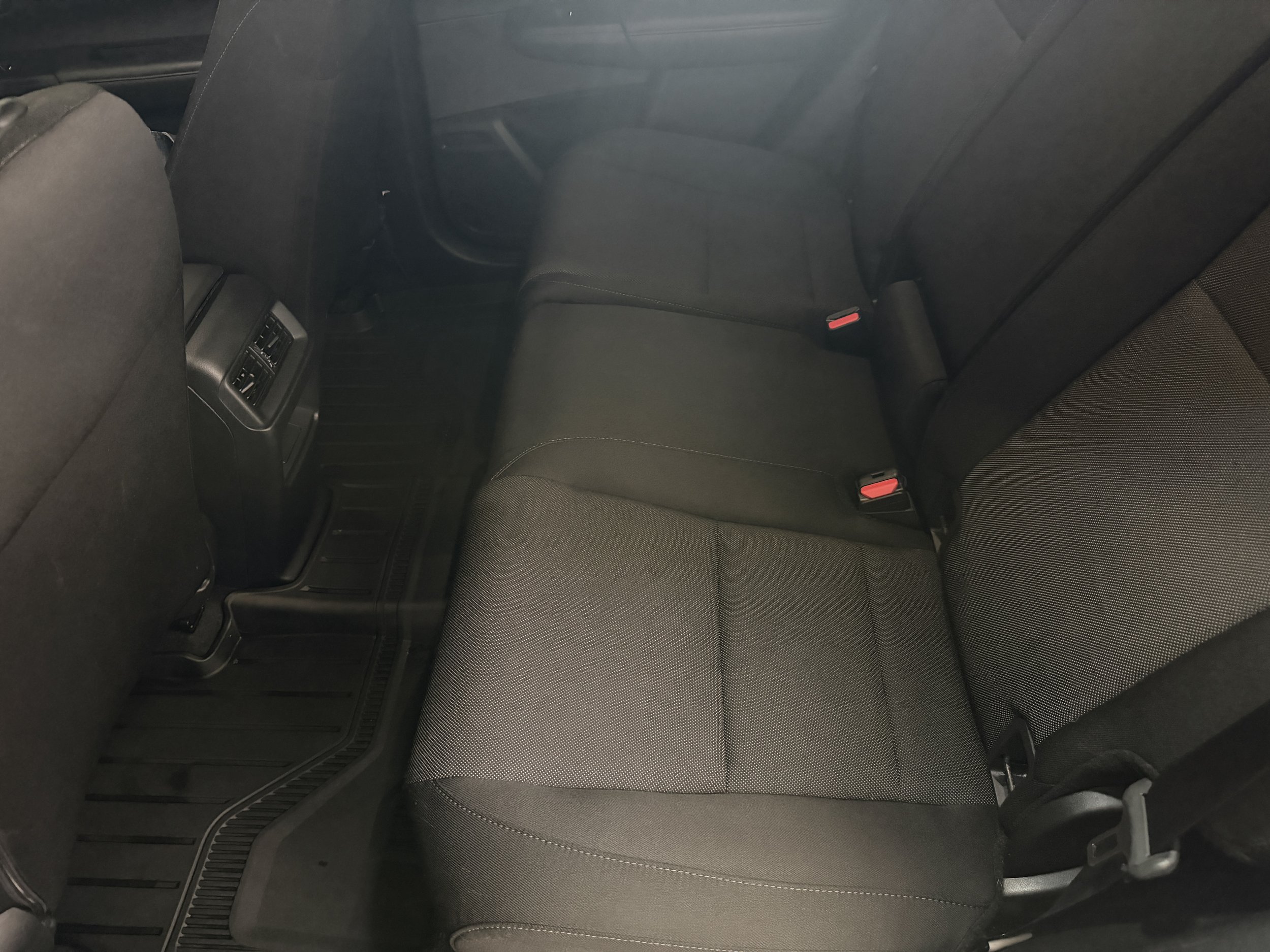The backseat of a car showing two black fabric seats with visible seat belts, and the rear climate control vents in the center console.