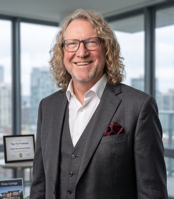 A smiling man with curly gray hair, glasses, and a beard, wearing a gray suit jacket, white shirt, and a maroon pocket square, standing in an office with large windows and city buildings in the background.
