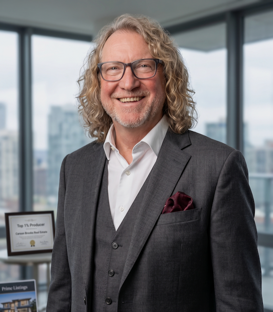 A smiling middle-aged man with curly blonde hair and glasses, dressed in a gray suit with a white shirt and a pocket square, standing in an office with large windows overlooking a cityscape.