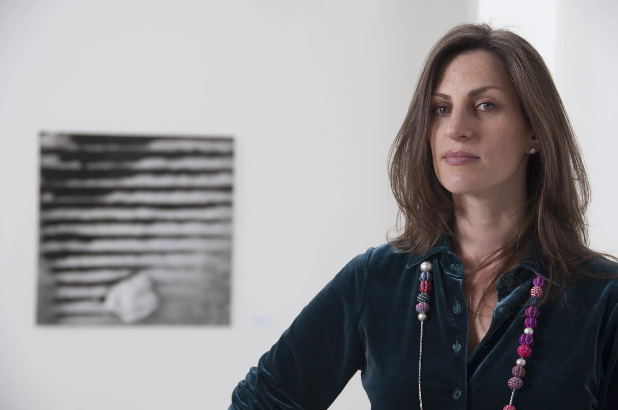 A woman with shoulder-length brown hair and a serious expression wearing a dark teal velvet shirt and colorful beaded necklaces stands near an art piece on a white wall.