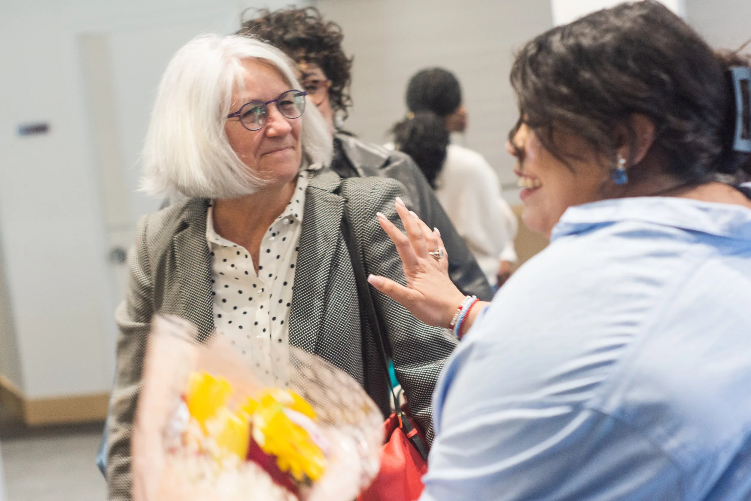 Two women, one with white hair and glasses, and the other with short dark hair, are engaged in conversation at an indoor event. Several other people are visible in the background.