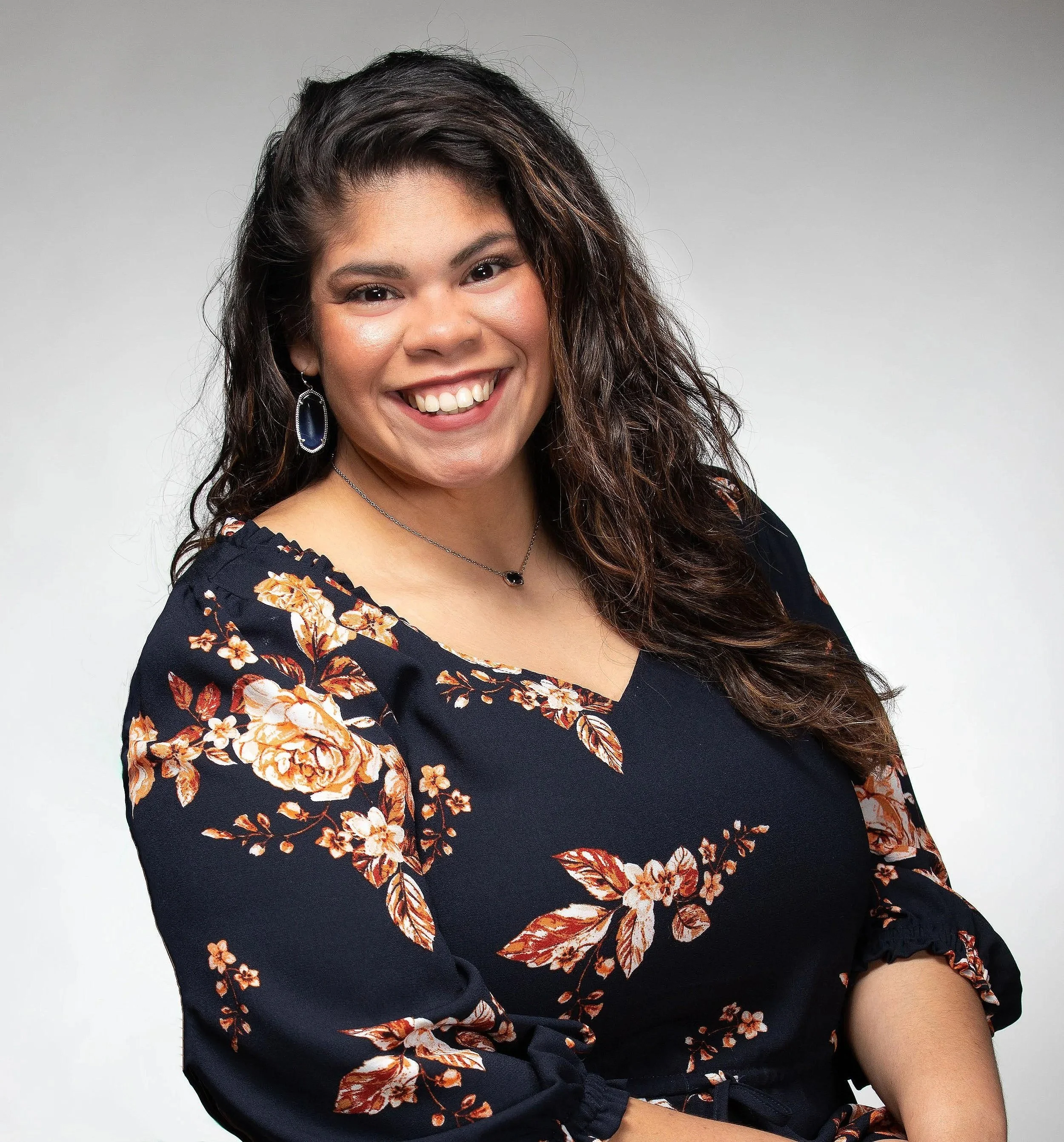 Portrait of a woman with long wavy dark hair, smiling, wearing large earrings, a necklace, and a floral-patterned black top against a gray background.