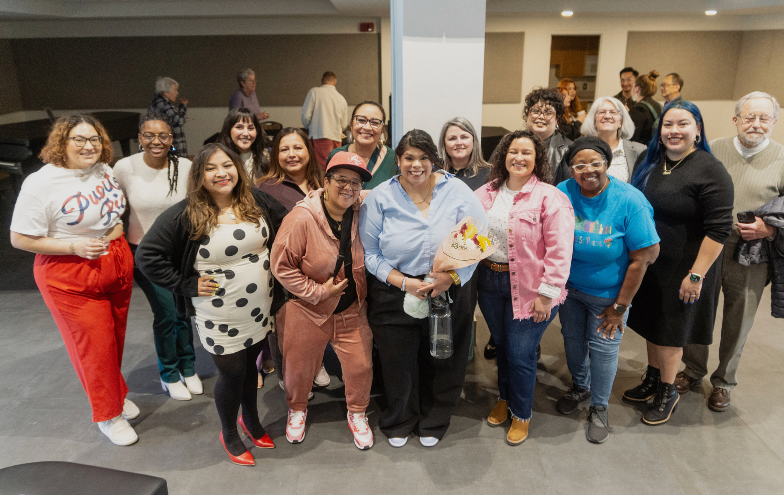 Group of diverse women smiling and posing together in an indoor setting, some holding flowers and a water bottle.