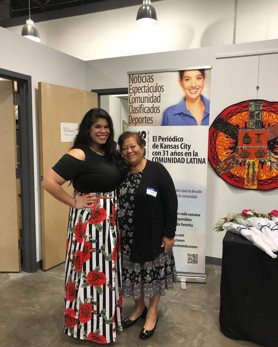 Two women standing together indoors, smiling. The woman on the left has long dark hair and wears a black top with a long striped skirt with red roses. The woman on the right has shorter hair and wears a black cardigan over a patterned dress. Behind them is a white wall with posters and artwork, including a poster with a smiling woman, and a colorful circular artwork on the right.