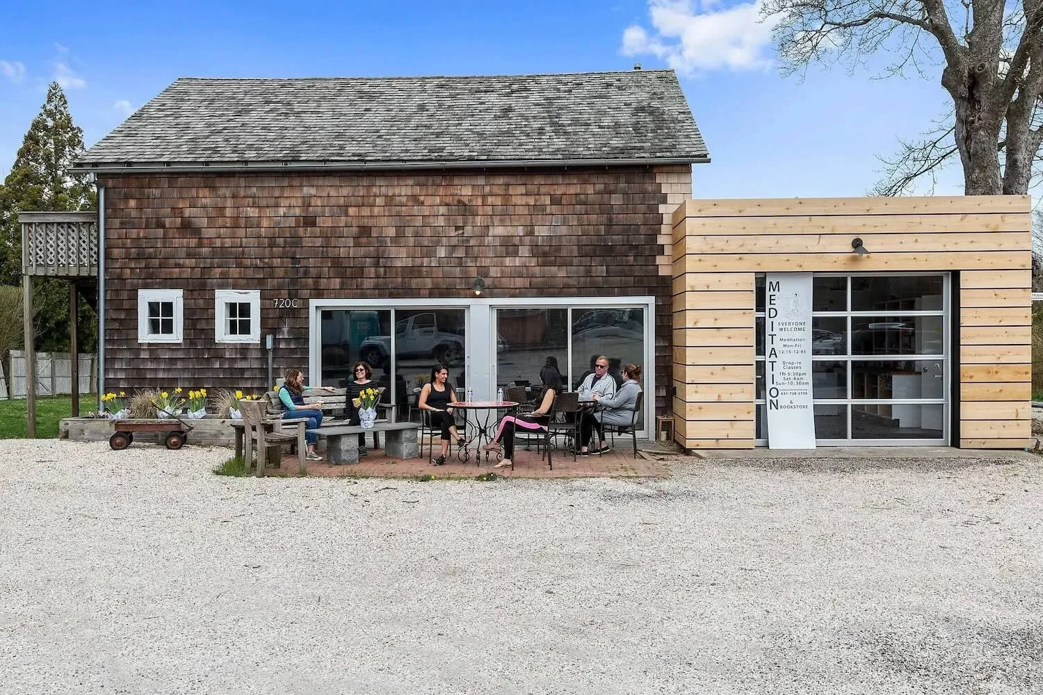 People sitting at outdoor tables in front of a building with wood and shingle exterior, offering meditation classes and a bookstore, with trees and a blue sky in the background.
