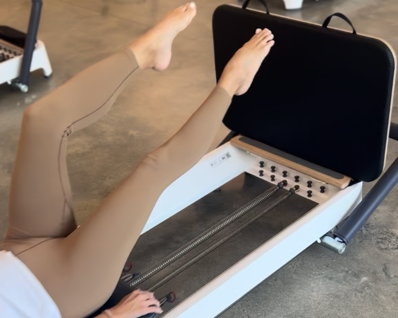 A woman exercise on reformer Pilates machine in a gym with dark walls and windows.
