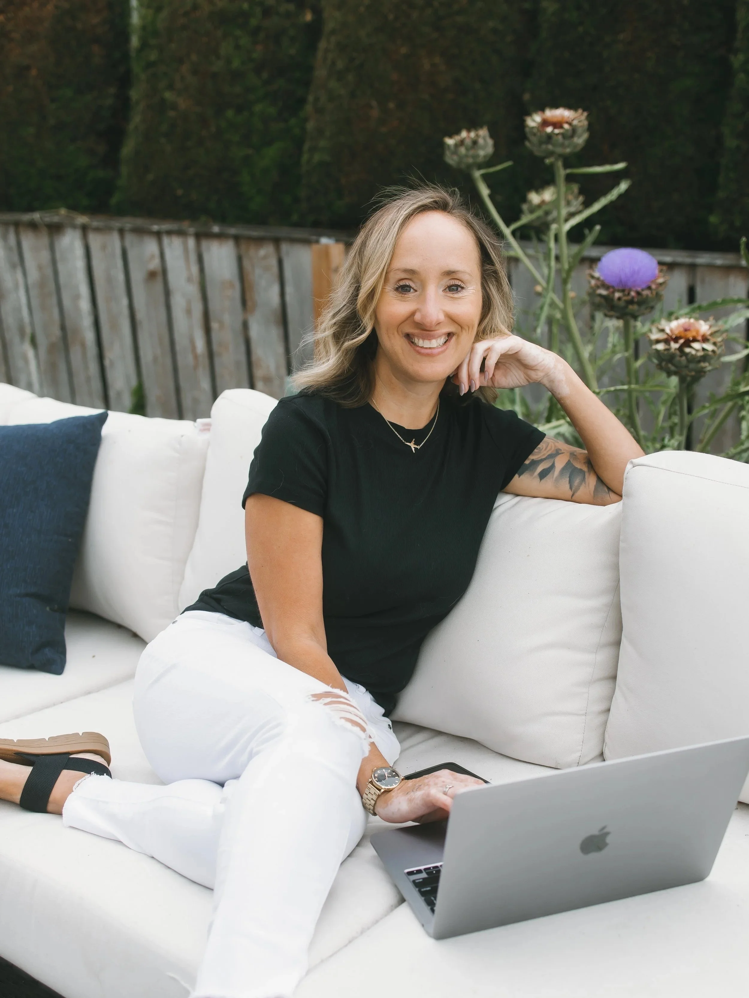 A woman with blonde hair, wearing a black t-shirt and white ripped jeans, sitting on a white outdoor sofa with pillows, smiling at the camera, with a laptop on her lap and a cellphone nearby, outdoors with plants and a wooden fence in the background.