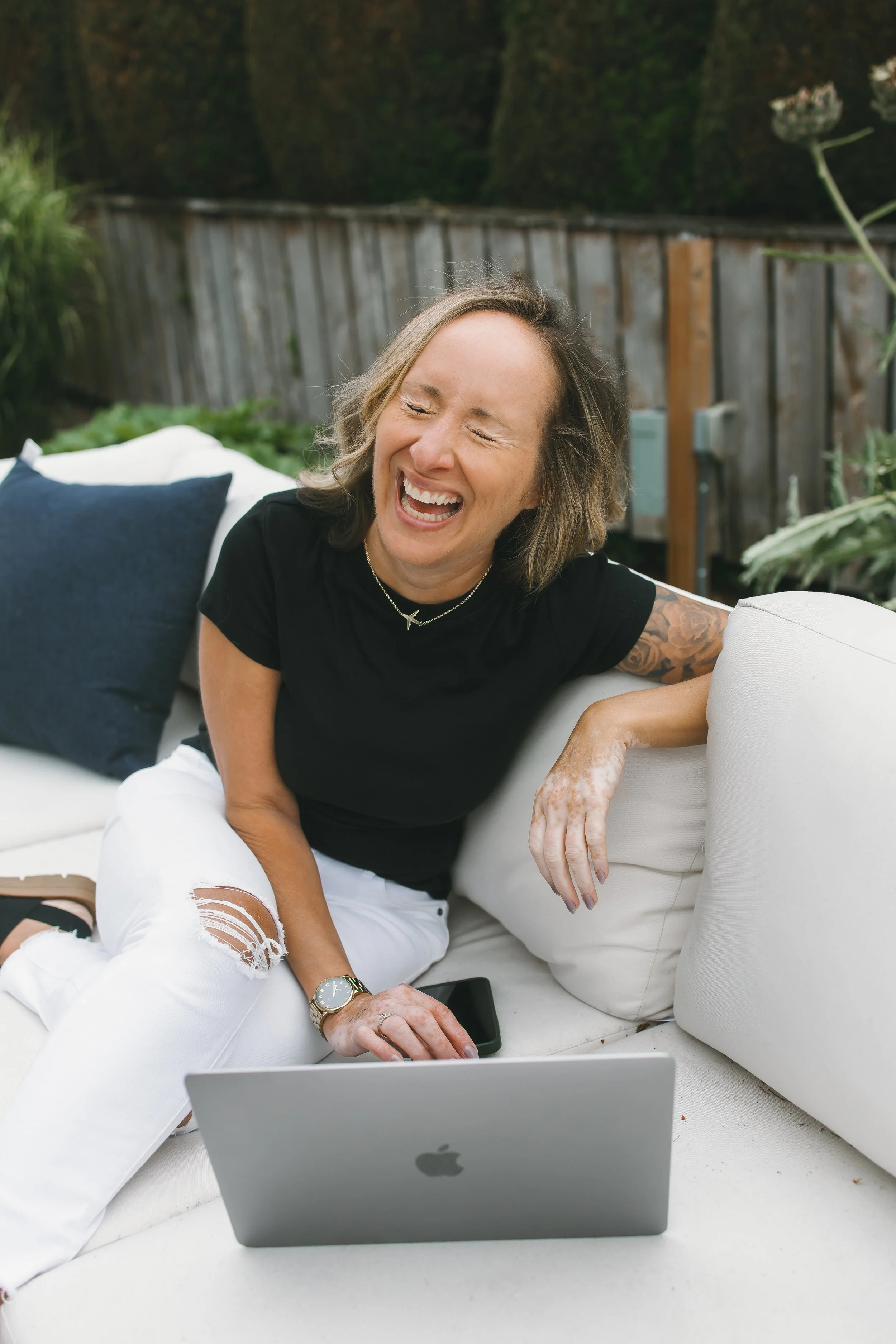 Woman with short blonde hair laughing and sitting on an outdoor couch with a laptop and smartphone, wearing a black shirt and white ripped jeans, in a backyard setting.
