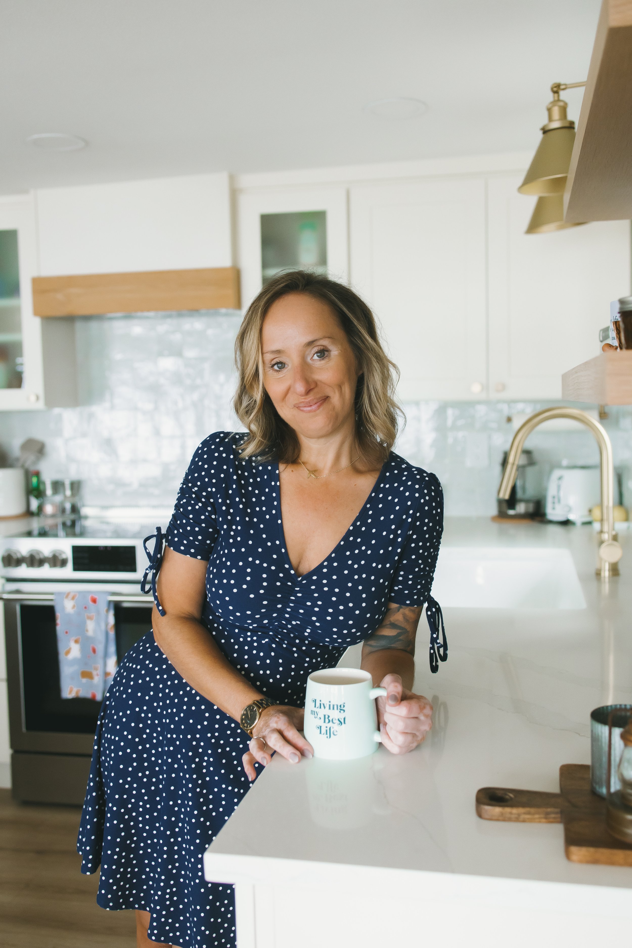 A woman with blonde hair, wearing a navy blue polka dot dress, stands in a modern kitchen holding a white mug with the words "Living my Best Life" printed on it. The kitchen has white cabinets, a stainless steel stove, and a countertop with a wooden cutting board and jars.