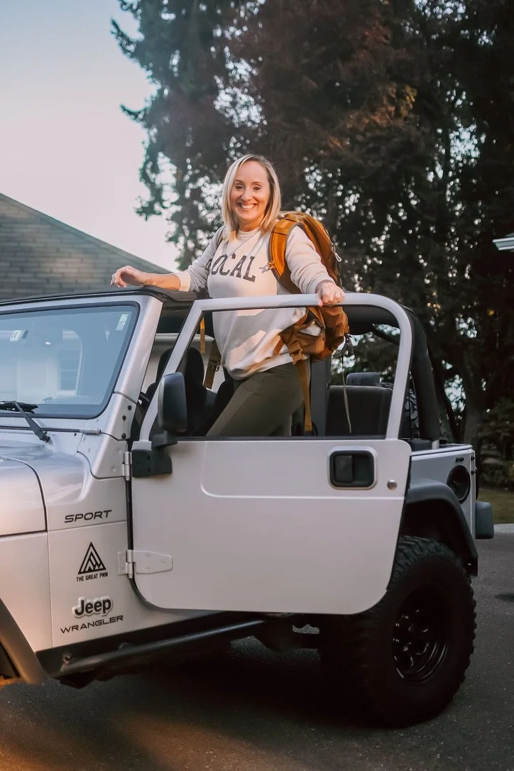 A smiling woman with a backpack standing in an open-top Jeep Wrangler, park in a residential neighborhood during dusk.