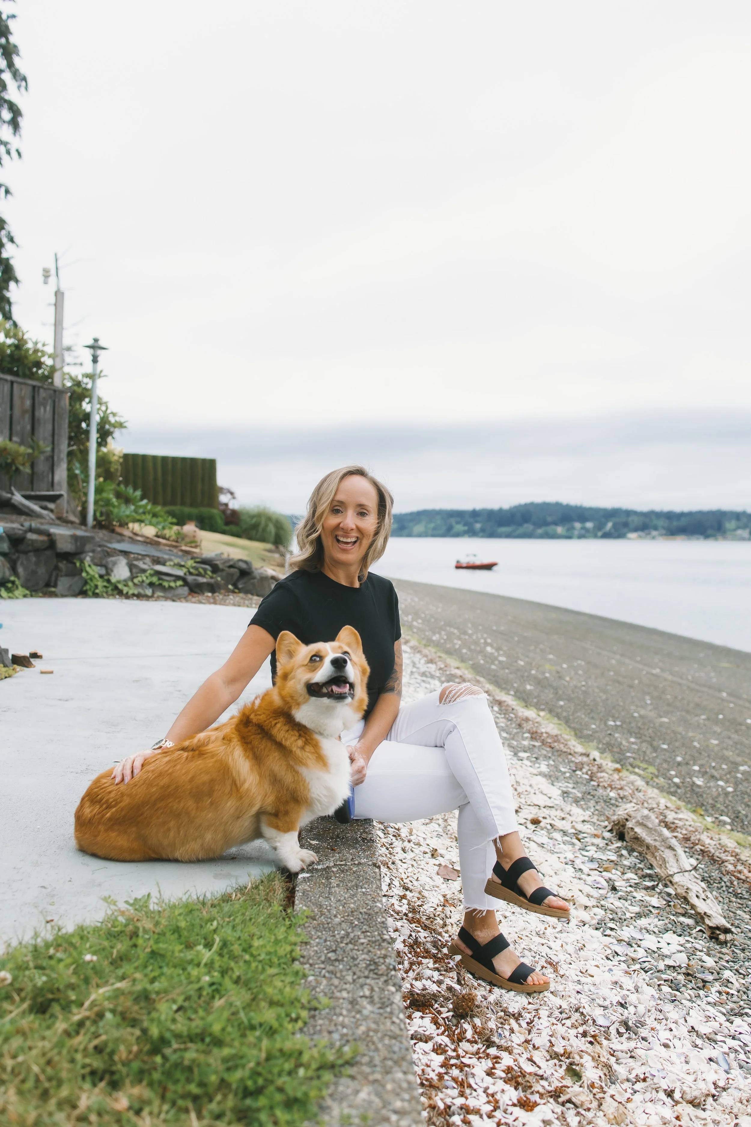 A woman with blonde hair, wearing a black top and white pants, sitting on a concrete curb by a lakeshore with her dog, a Pembroke Welsh Corgi, beside her. The background shows water, a boat, and a distant shoreline under cloudy skies.