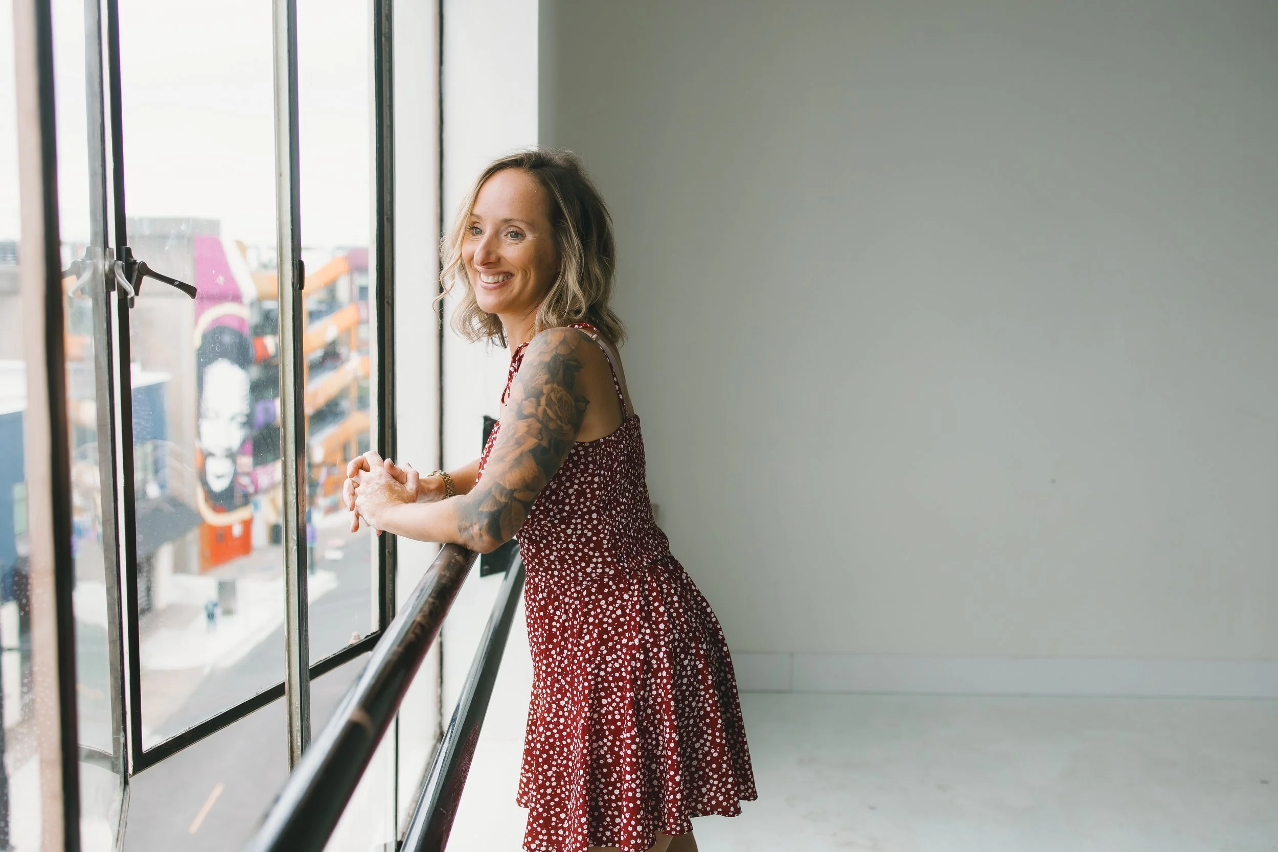 A woman in a red dress with white polka dots is standing at a large window, smiling, with greenery and buildings visible outside.