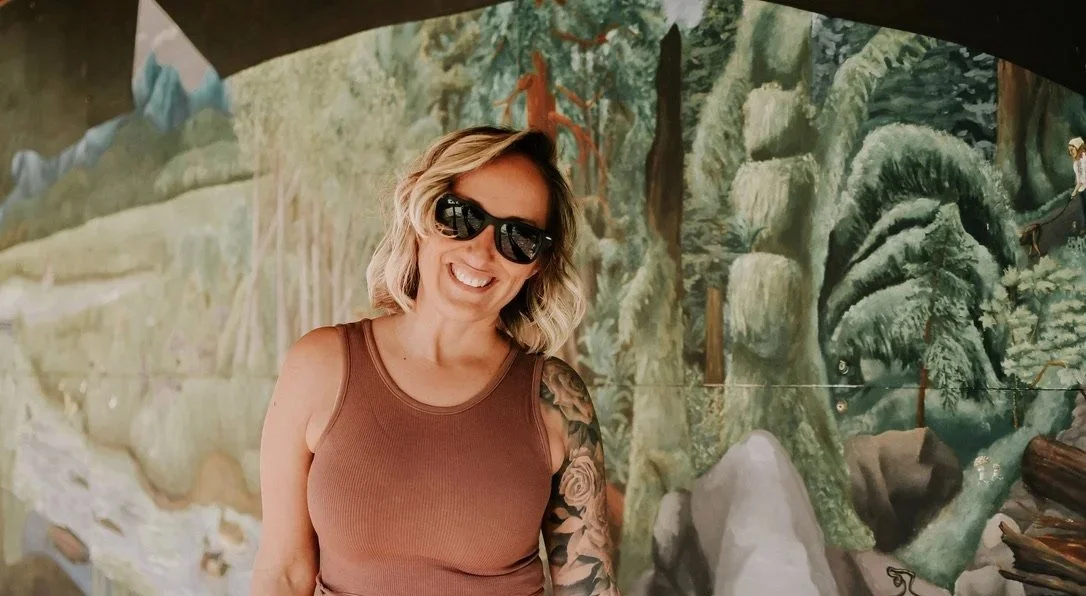 A woman with blonde hair, wearing sunglasses and a brown tank top, smiling in front of a painted forest mural.