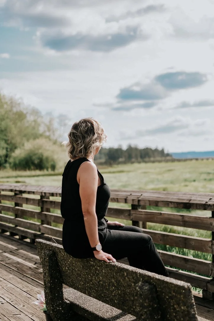A woman sitting on a wooden bench on a bridge overlooking a green field and trees, with a cloudy sky overhead.