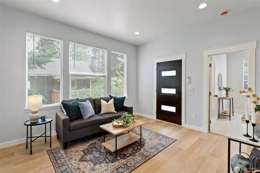 Modern living room with a dark front door, a gray sofa with pillows, a wooden coffee table, a black side table with a lamp, and large windows with blinds, overlooking a backyard.