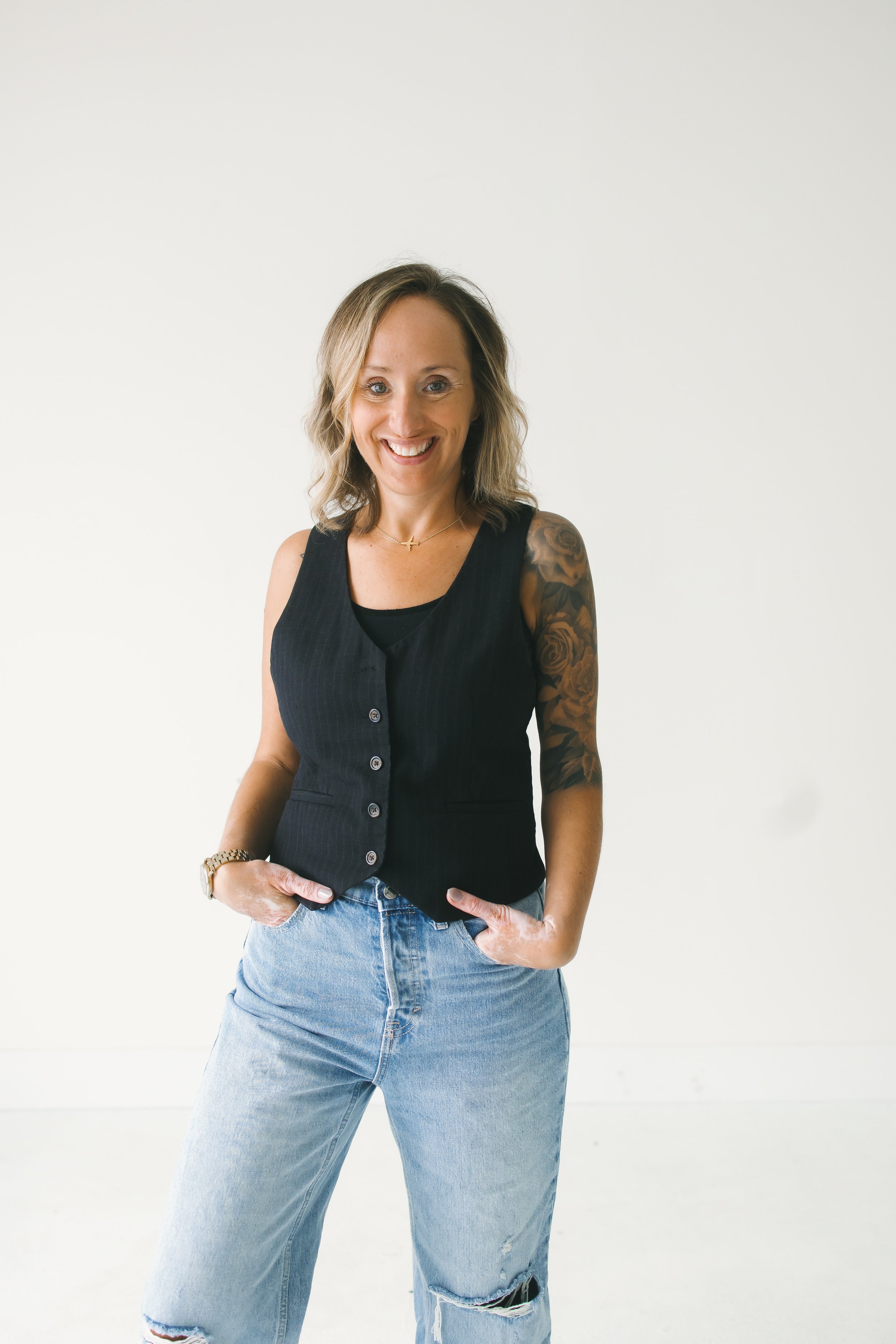 A woman smiling with her hands in her pockets, wearing a sleeveless black button-up top and light blue ripped jeans, standing against a plain white background.