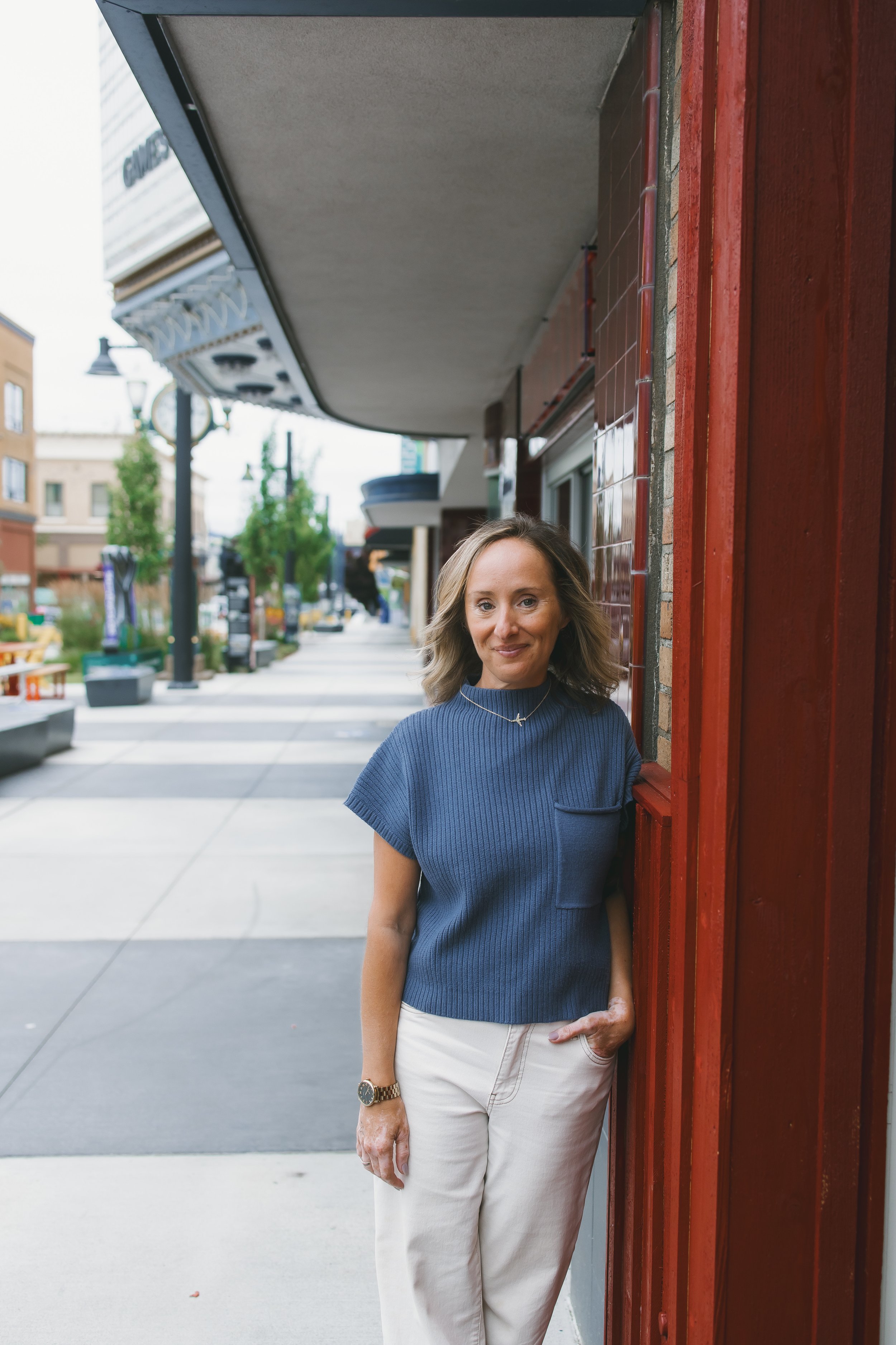 A woman with wavy blonde hair, wearing a blue ribbed short-sleeve top, white pants, a wristwatch, and a delicate necklace, stands with her hands in her pockets against a red wooden wall on a city sidewalk, smiling at the camera.