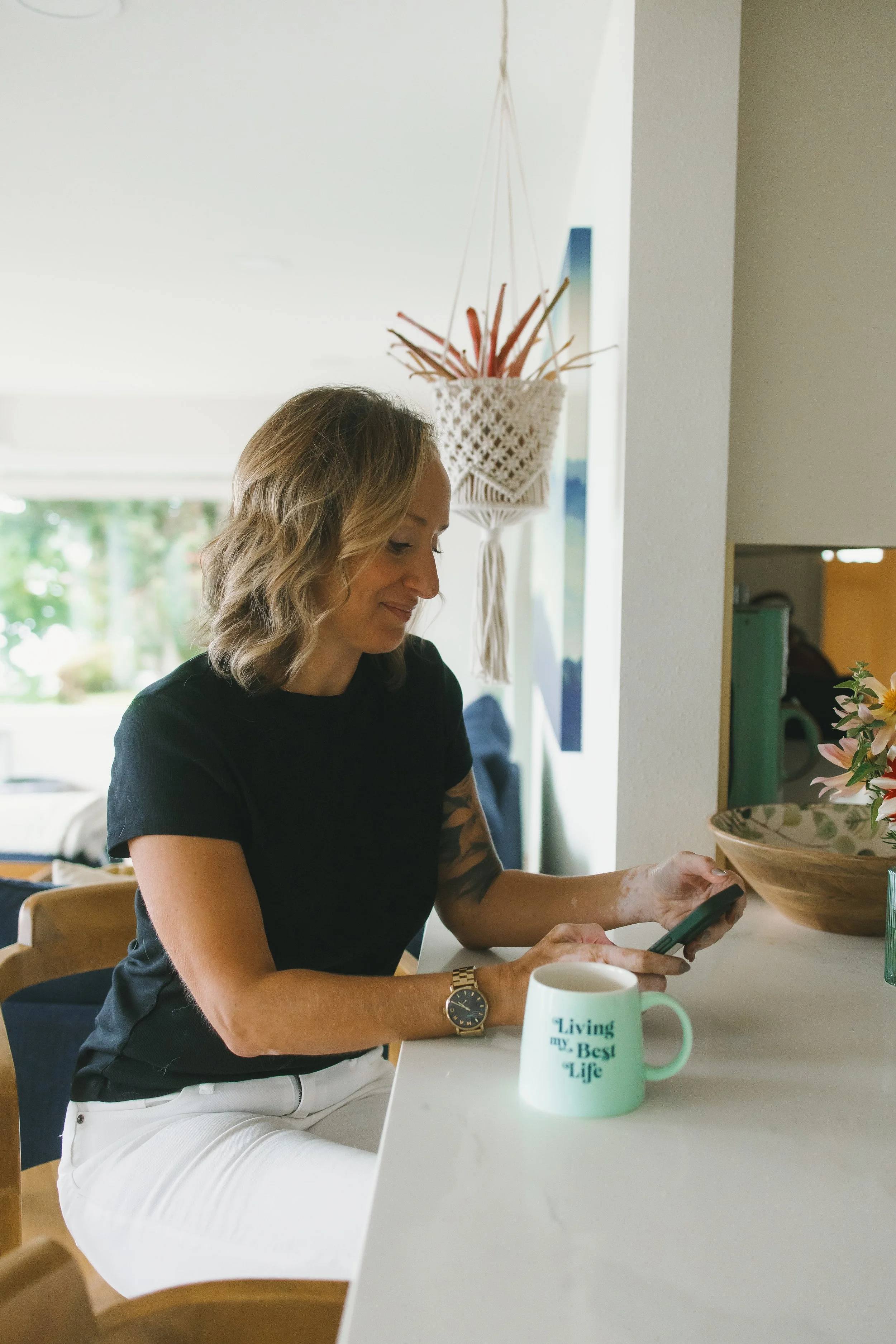 A woman with wavy blonde hair, wearing a black shirt and white pants, sitting at a table with a mug that reads 'Living my Best Life,' holding a smartphone. In the background, there is a wall hanging and a room with greenery visible through a window.