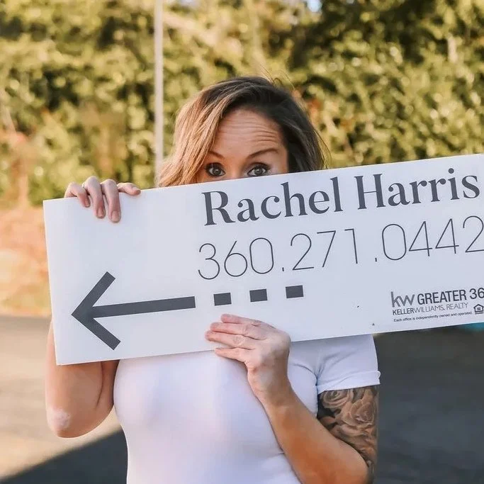 Woman holding a large sign with her right eye peeking over it, outdoors with trees in the background.