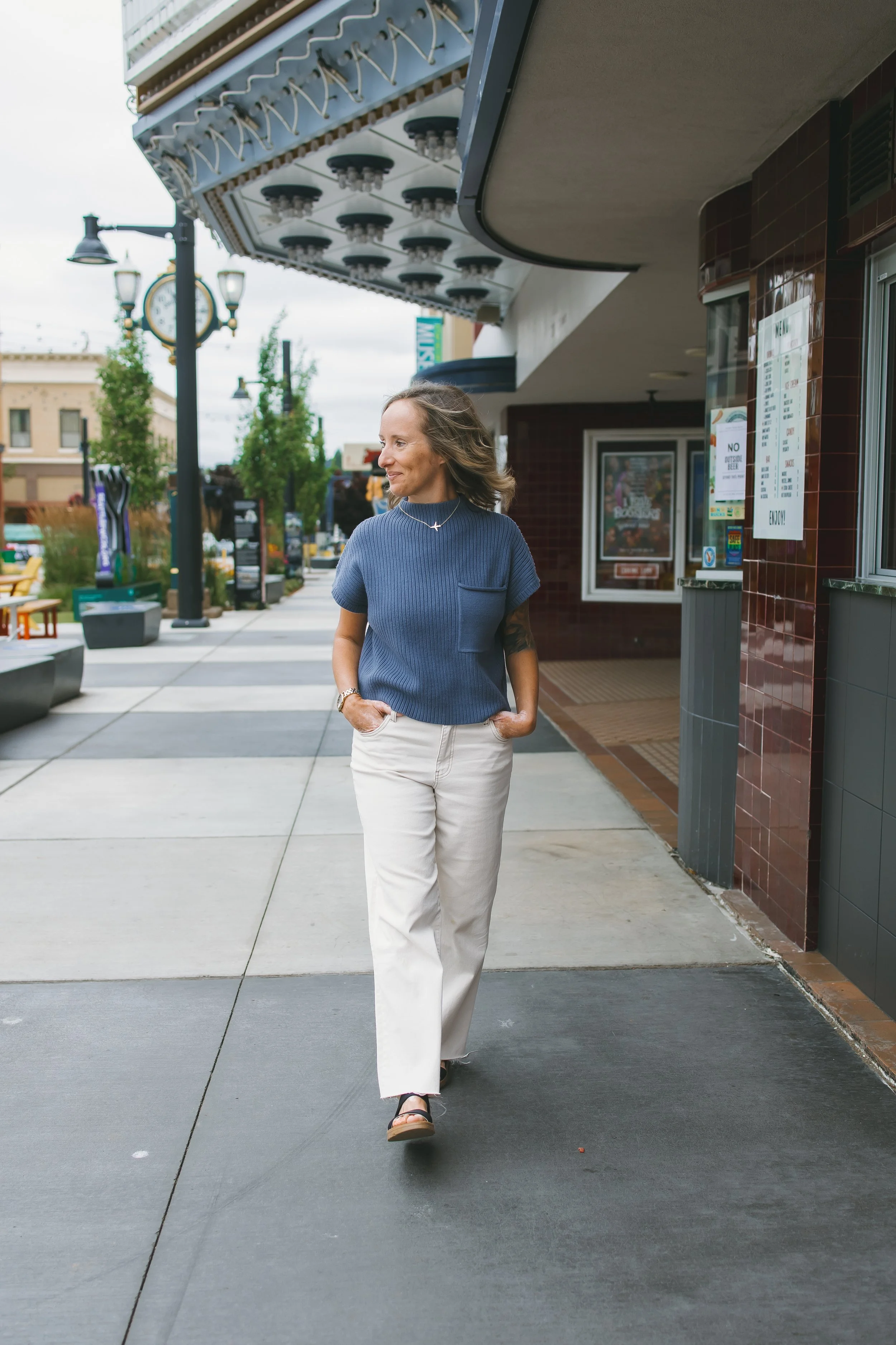 A woman walking on a sidewalk outside a building with a movie theater marquee. She is wearing a blue sweater, white pants, sandals, and has her hands in her pockets, with her hair flowing in the breeze.