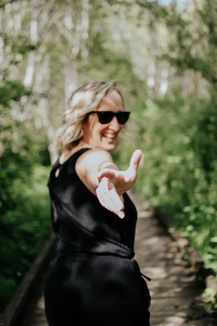A woman with blonde hair, wearing sunglasses and a black outfit, smiling and extending her hand towards the camera on a wooden bridge in a lush green outdoor setting.