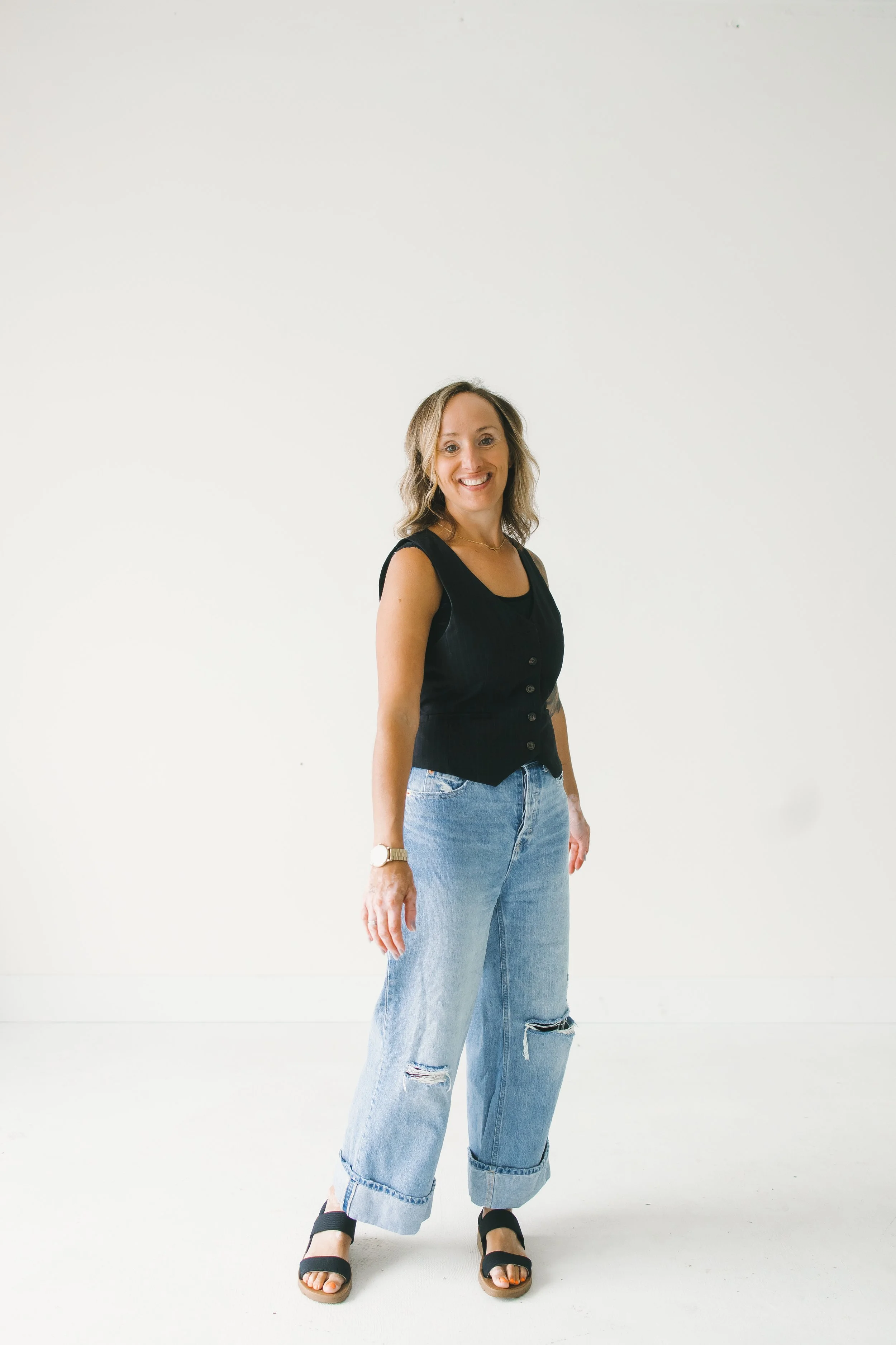 A woman with blonde hair smiling, wearing a black sleeveless top, ripped jeans, black sandals, and a watch standing against a plain white background.