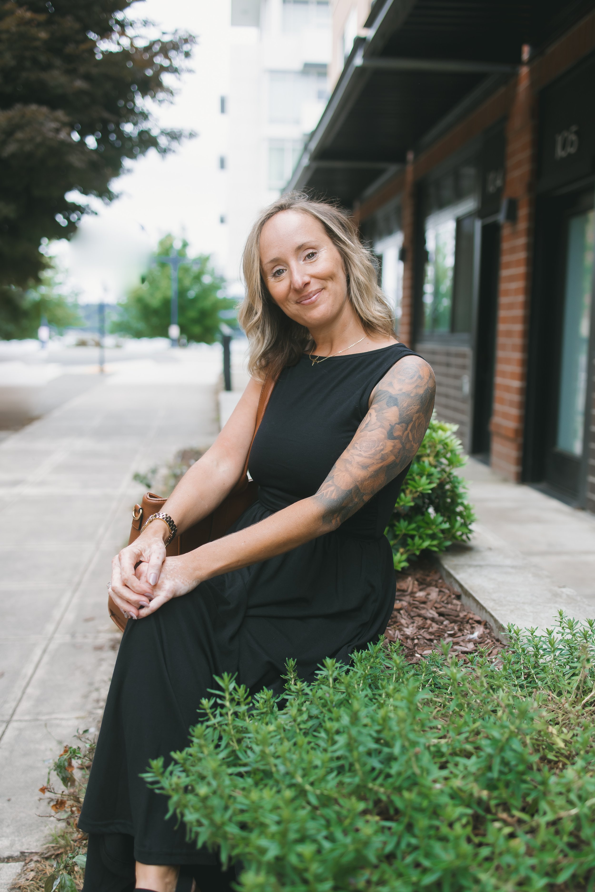A woman with wavy blonde hair, wearing a black sleeveless dress, sits on a bench beside a sidewalk lined with green bushes and trees in an urban area, smiling at the camera.