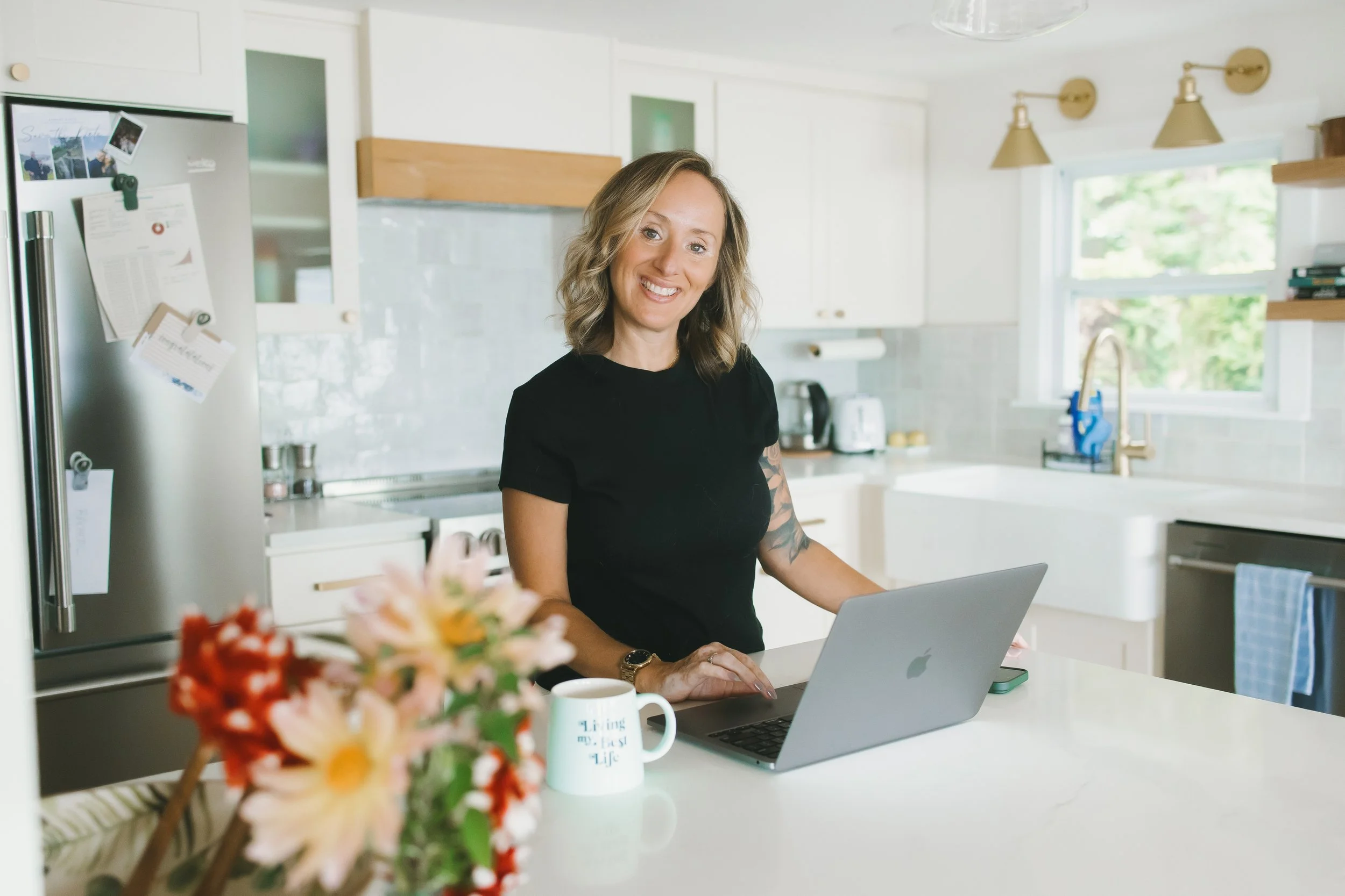 A woman with shoulder-length wavy blonde hair, wearing a black shirt, sitting at a kitchen counter working on a laptop, with flowers and mugs on the counter, and a bright kitchen with white cabinets, a stainless steel refrigerator, and a window.