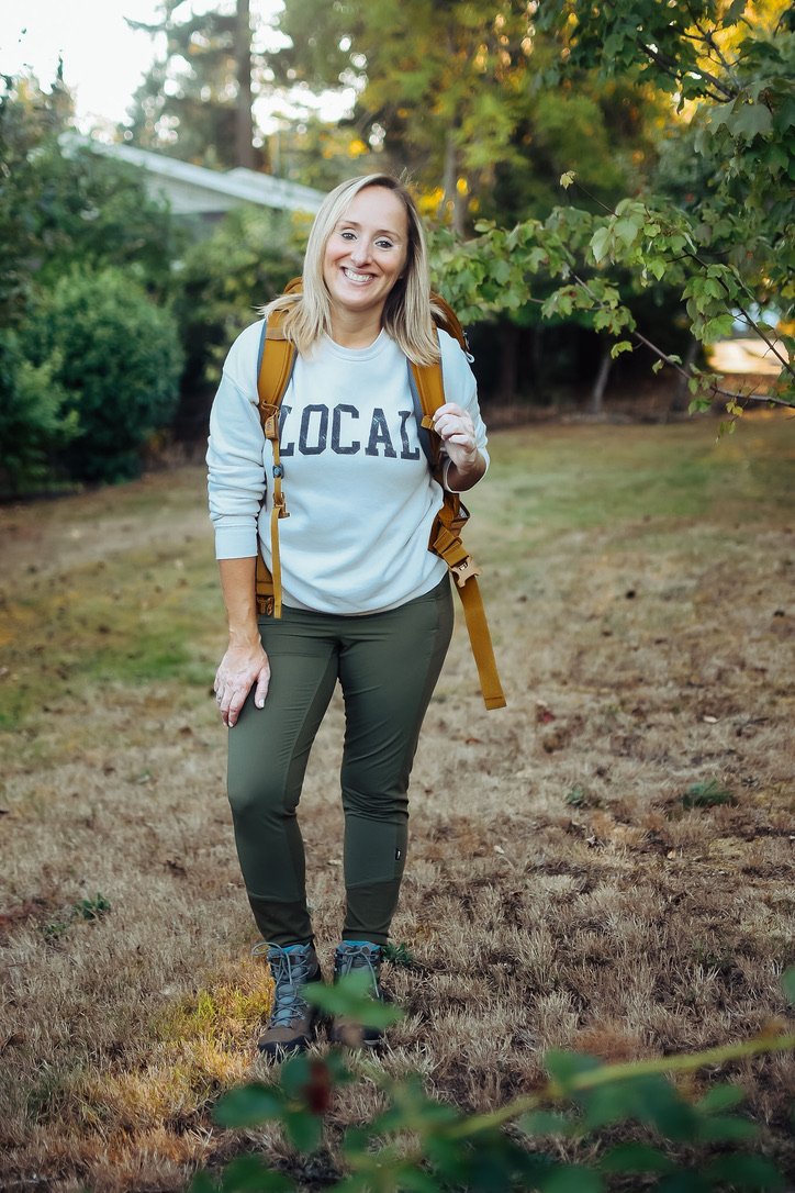 A woman with blonde hair smiling outdoors, wearing a light blue sweatshirt that says 'LOCAL', green pants, hiking boots, and a yellow backpack, standing on a grassy trail surrounded by trees.