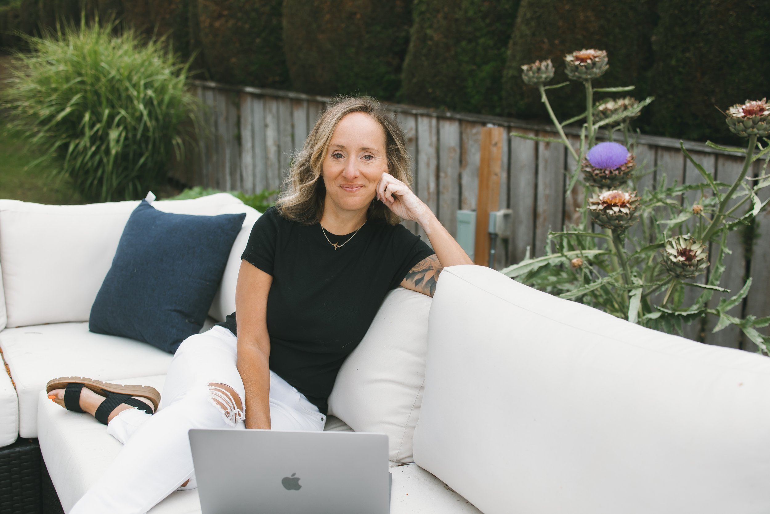 A woman sitting on a white outdoor sofa with a laptop in front of her, smiling at the camera. She has wavy, shoulder-length hair, wears a black t-shirt and white ripped jeans, and has a tattoo on her right arm. There are green plants, a wooden fence, and tall artichoke flowers in the background.