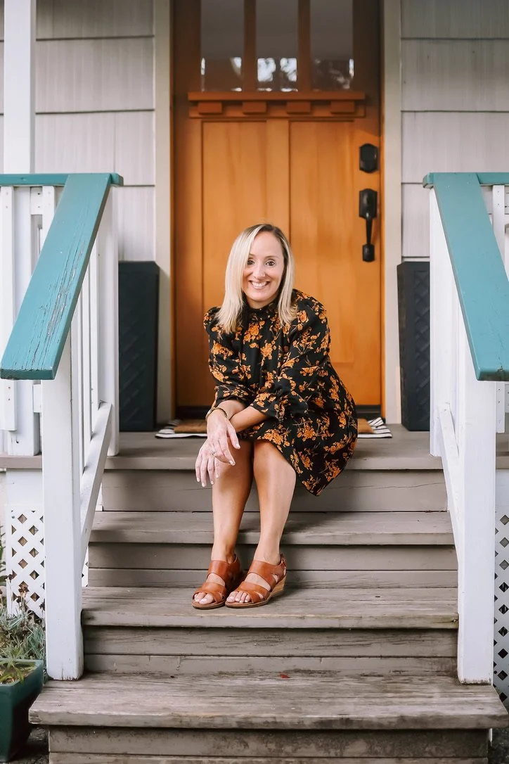 A woman in a black and orange floral dress sitting on the front steps of a house, smiling at the camera. The house has white siding, a wooden front door, and teal-painted handrails.