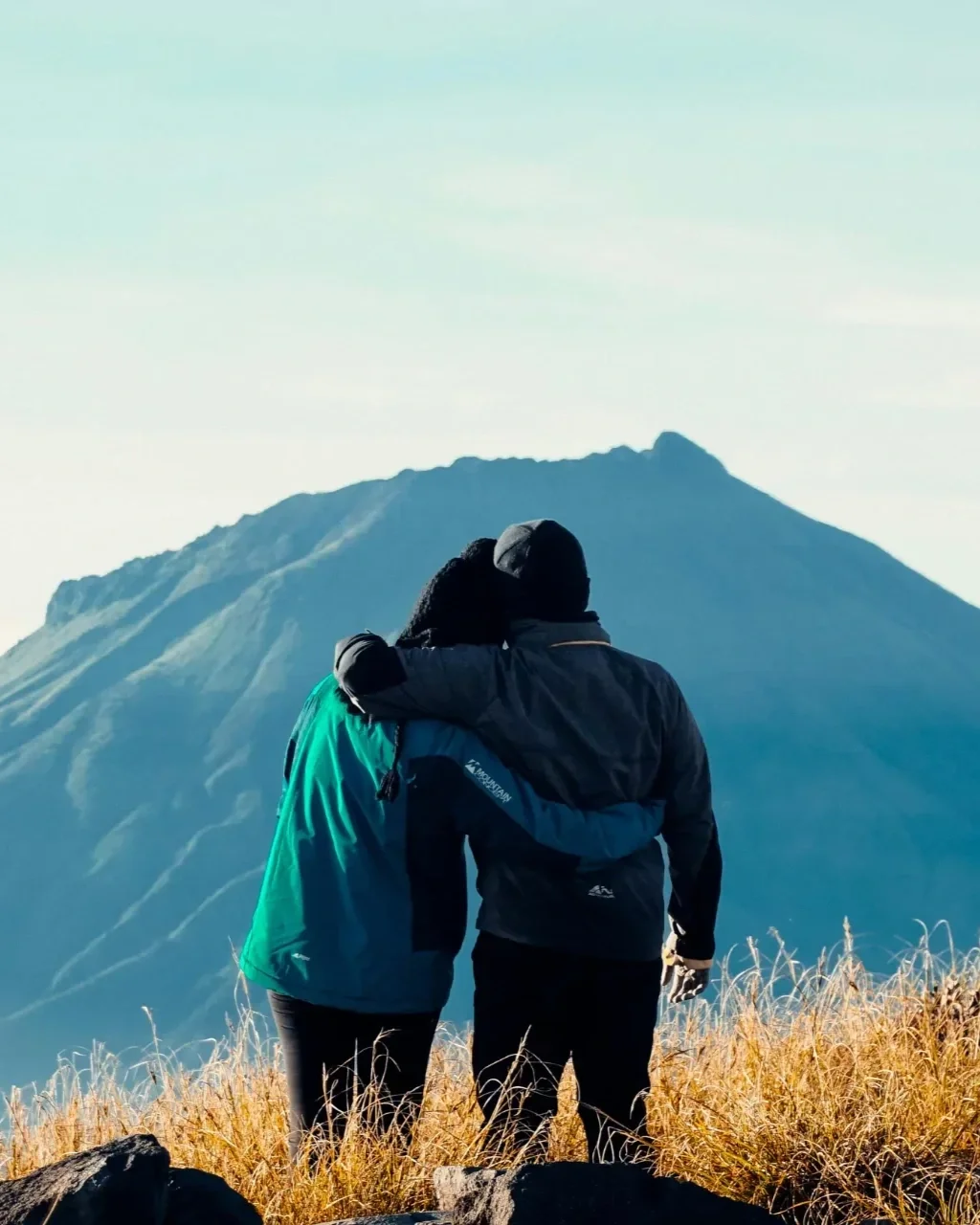A couple in warm clothing hugging on a grassy mountain overlooking a blue mountain landscape.