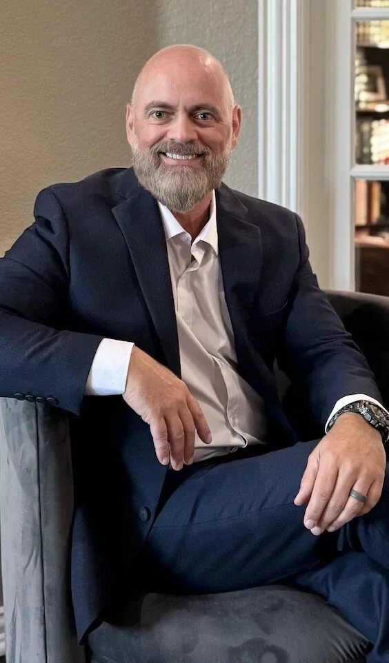 Dr. Russell Skinner, a middle-aged man, dressed in a dark suit and white shirt, sitting comfortably in a chair and smiling at the camera inside a room with beige walls and bookshelf.