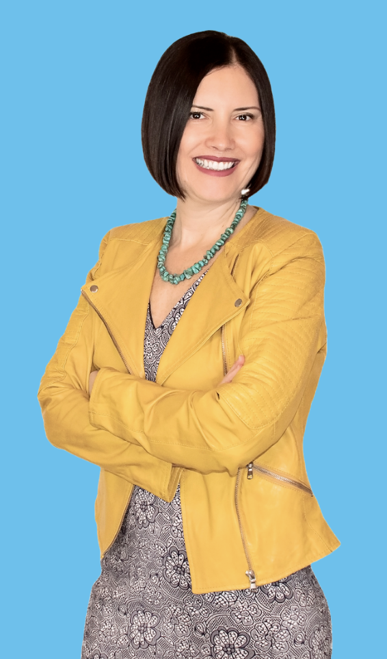 Dr. Ronit Mor, a woman with dark hair smiling, wearing a yellow leather jacket, a turquoise necklace, and a patterned dress, standing with arms crossed against a blue background.