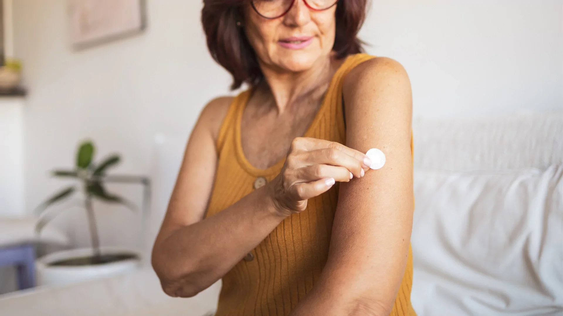An older woman with red hair and glasses is applying a white patch to her upper arm, possibly for vaccination or health measurement, in a cozy living room setting.