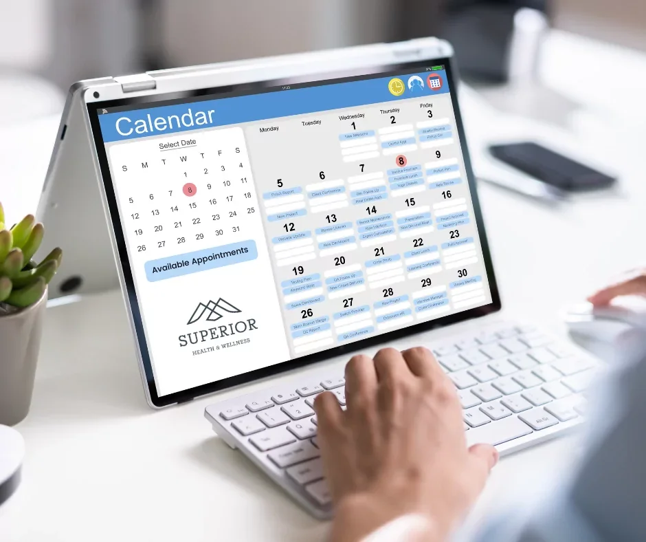 A tablet displaying a calendar for a health and wellness clinic, with appointments scheduled throughout the month, alongside a small potted succulent plant on the left and a person's hand typing on a white keyboard.